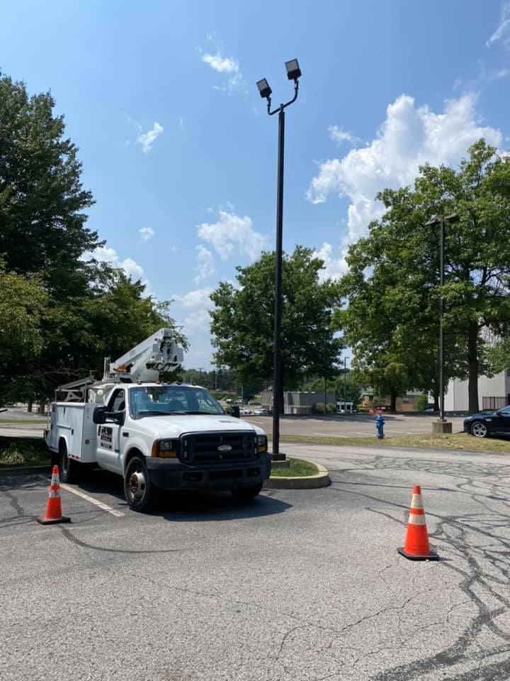 White utility truck parked near a tall light pole, cone in front. Sunny day.