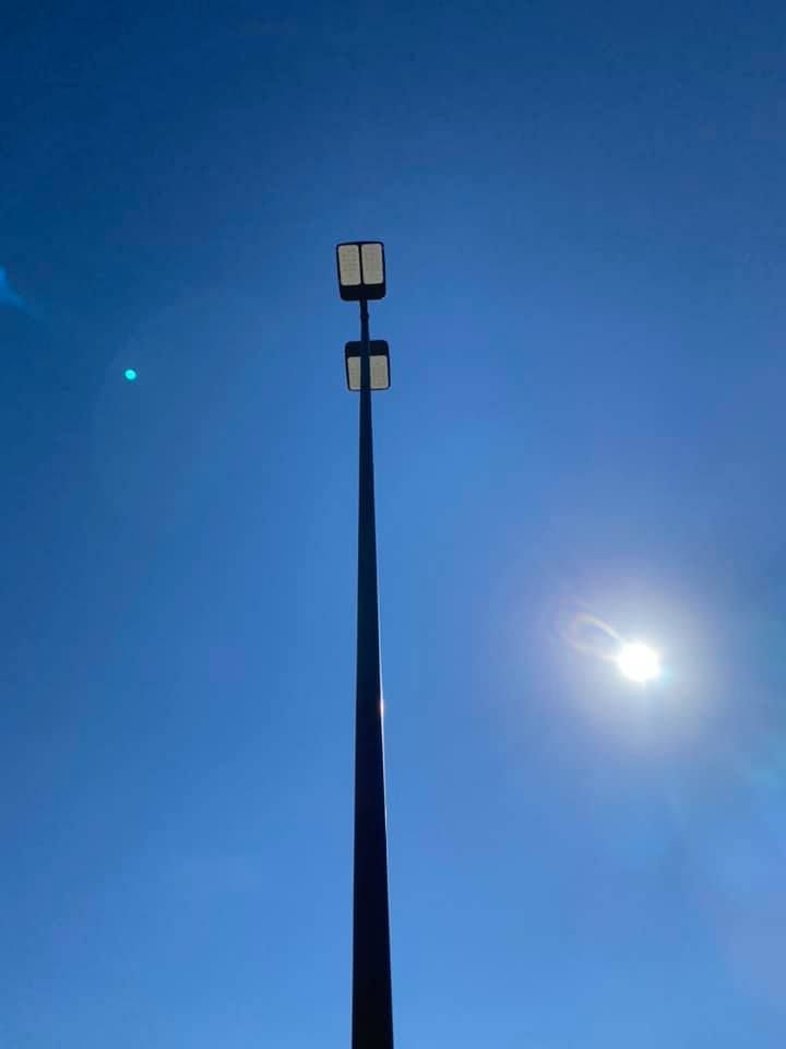 Tall black light pole with two rectangular lights against a bright blue sky with sun.