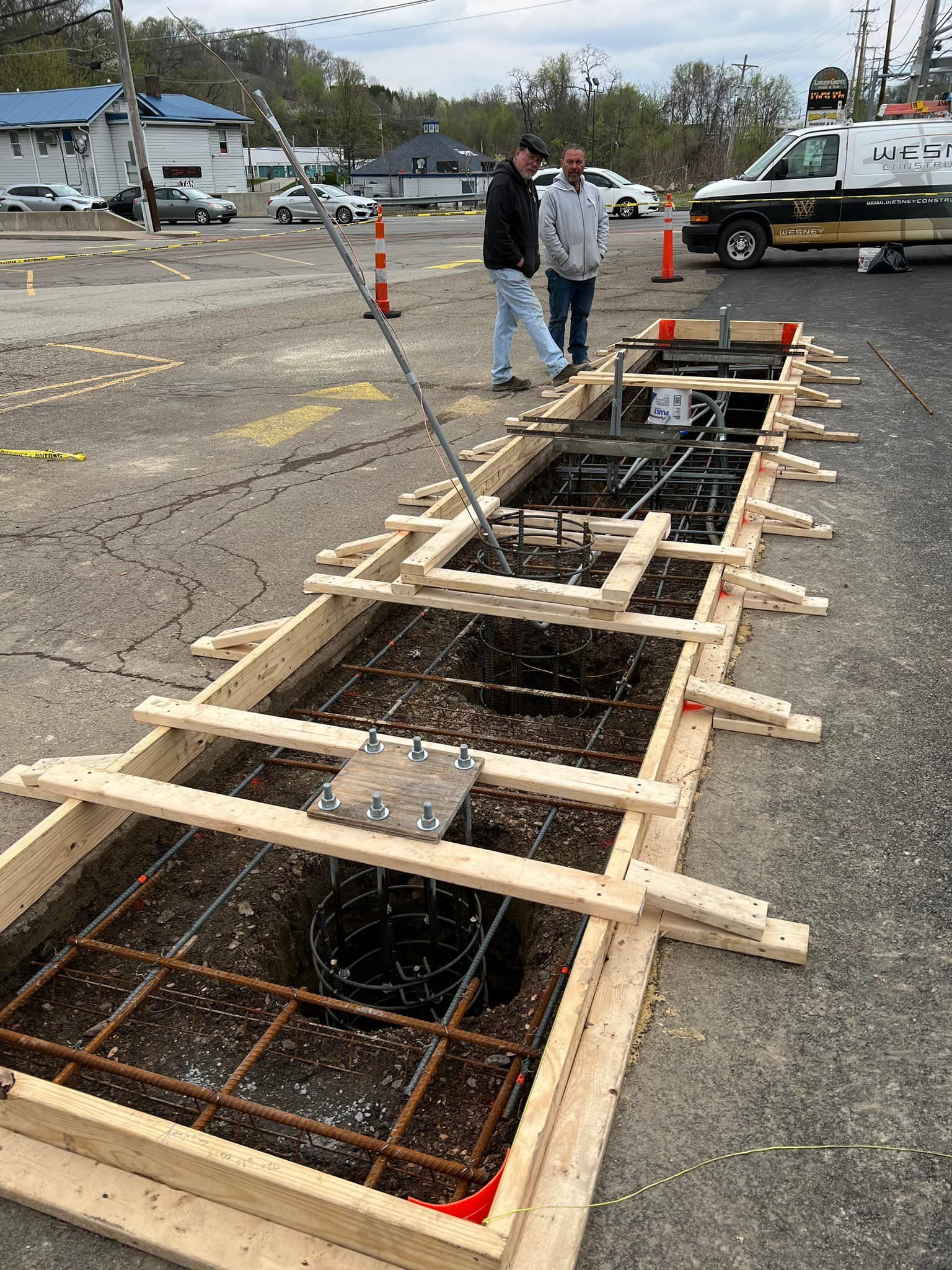 Construction site with wooden forms and metal reinforcement; two men observe the work.