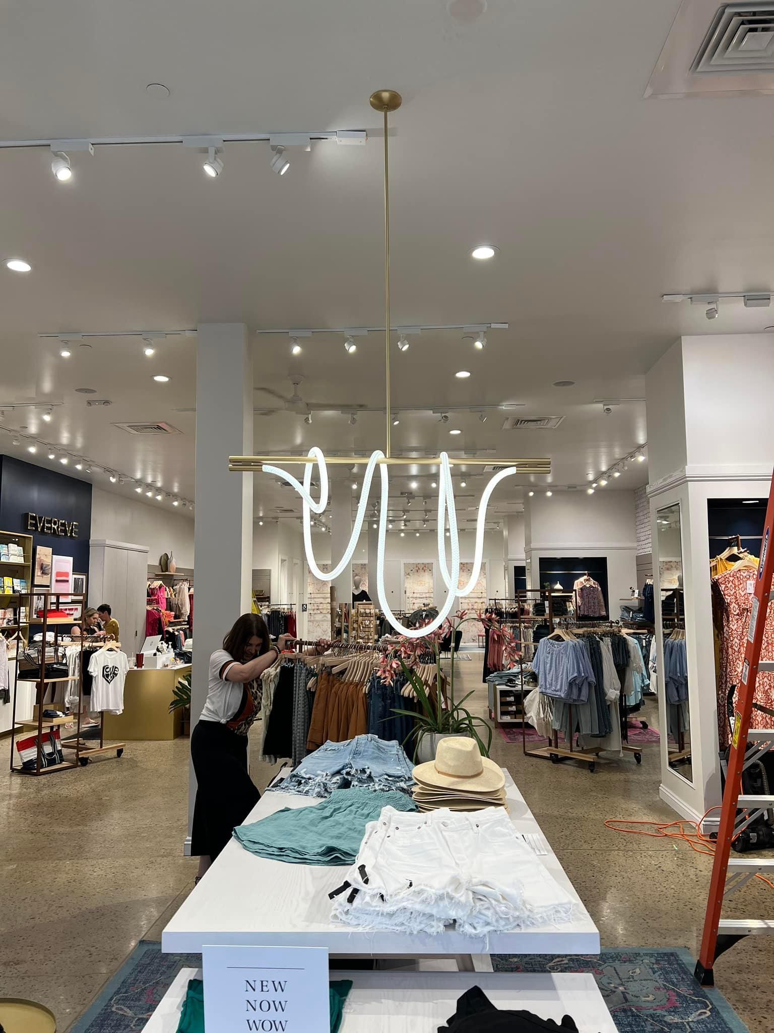 Woman adjusting a modern light fixture in a clothing store with merchandise on display.