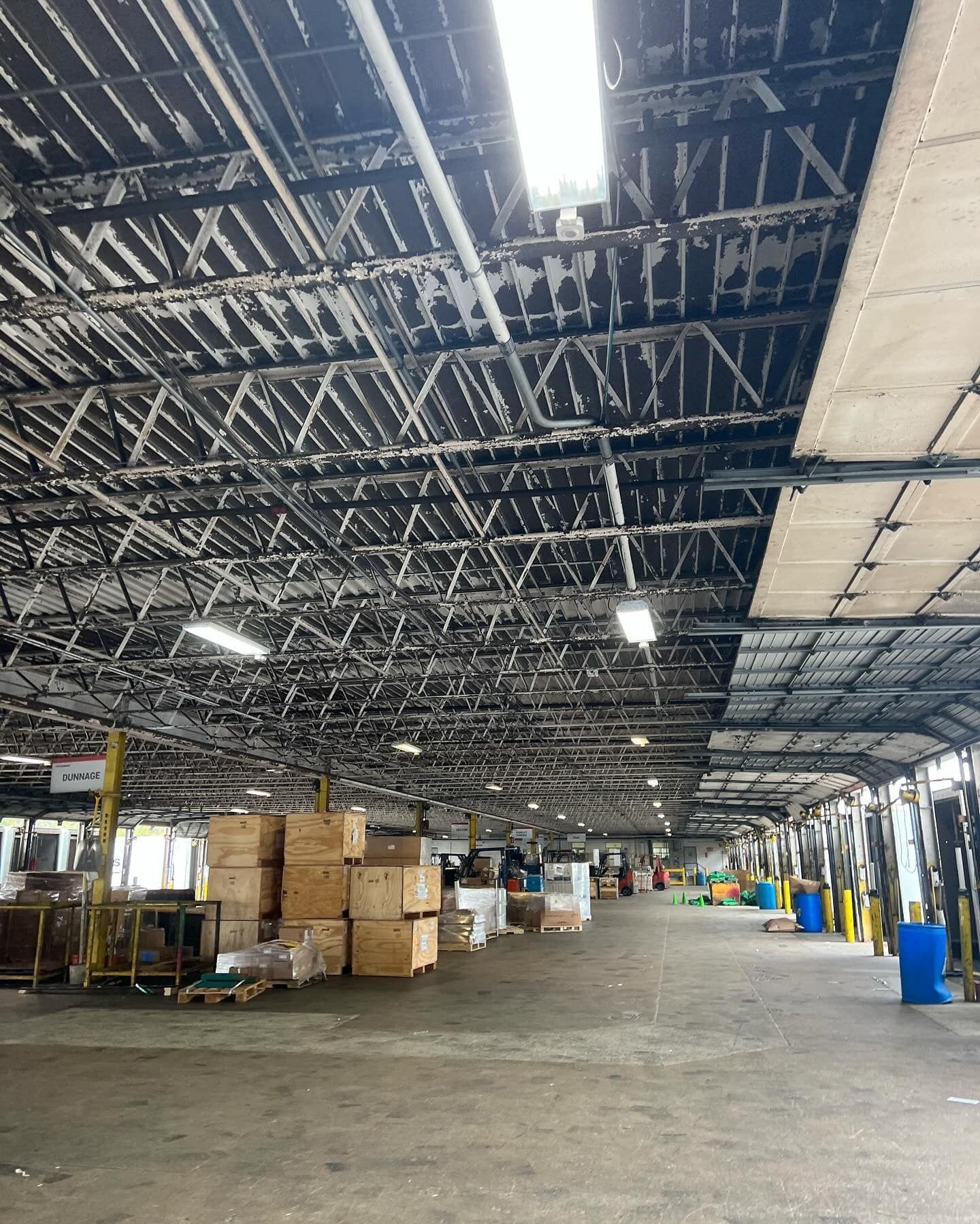 Warehouse interior with pallets, boxes, and a metal truss ceiling. Fluorescent lighting.