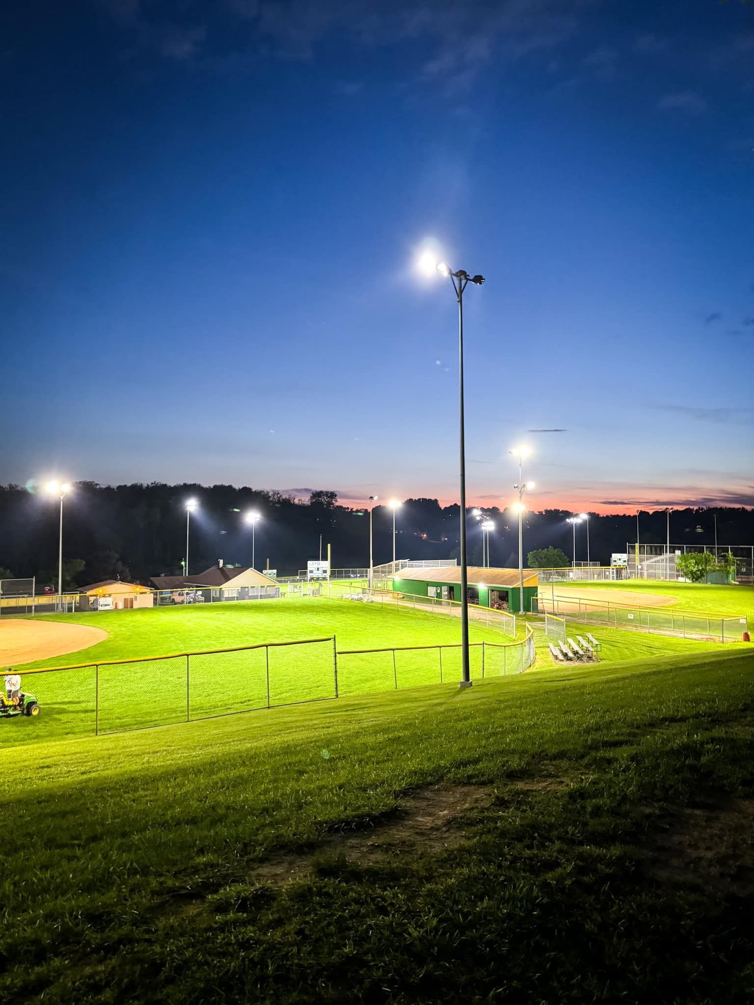 Baseball field at dusk, brightly lit by tall stadium lights against a blue and orange sky.