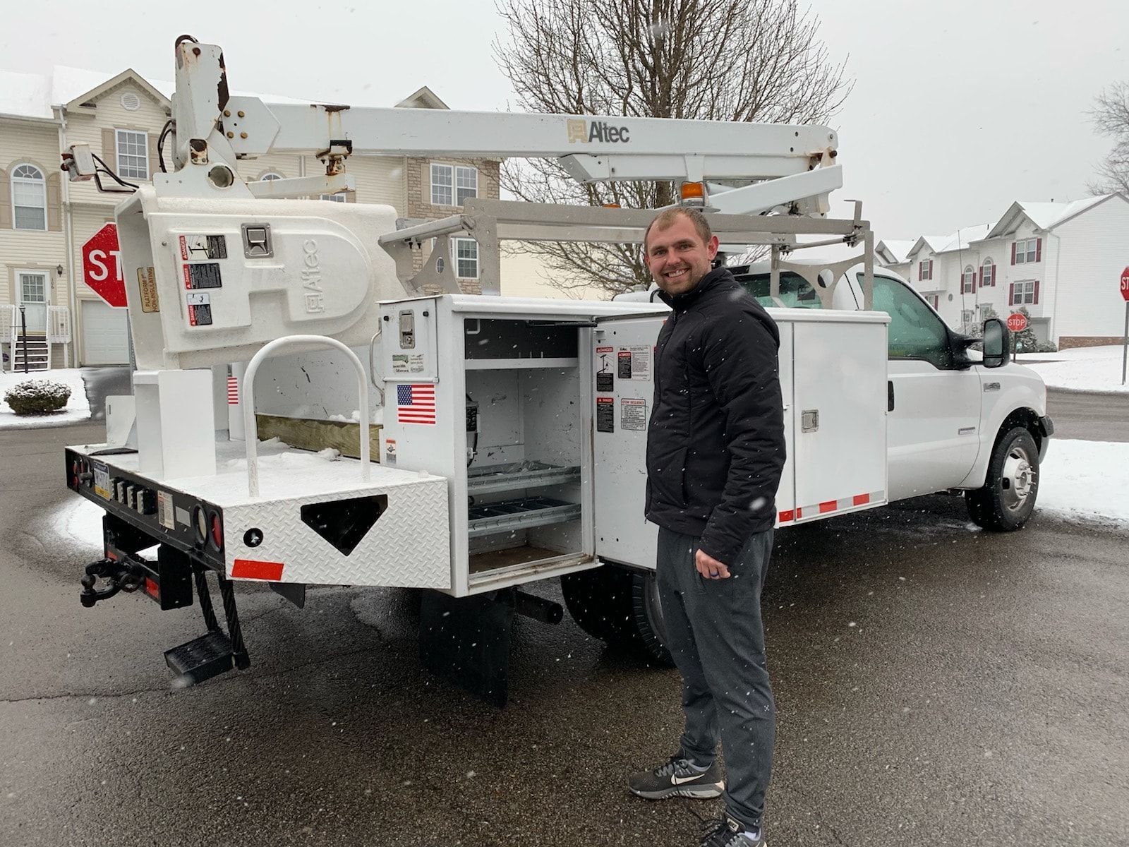 Man stands beside a white utility truck with an open tool compartment, with snow falling.