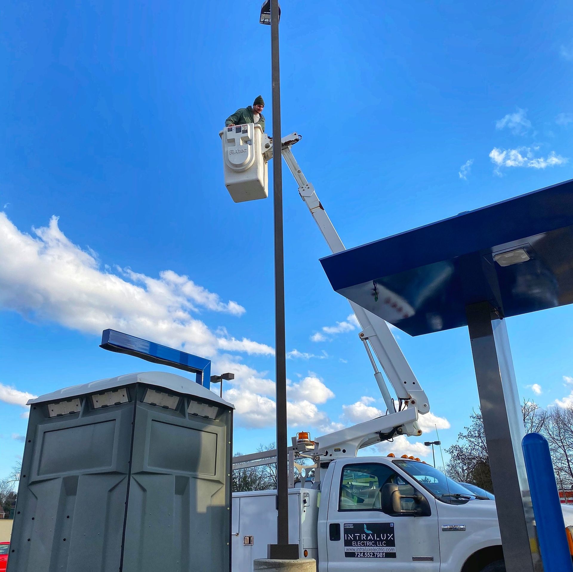 Person in a bucket lift repairing a tall light pole at a gas station under a blue sky.