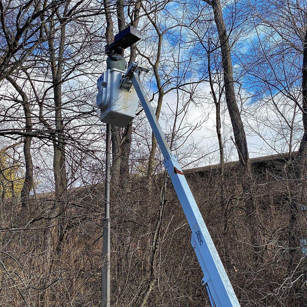 Bucket lift with a person working on equipment, against a backdrop of trees and a cloudy sky.