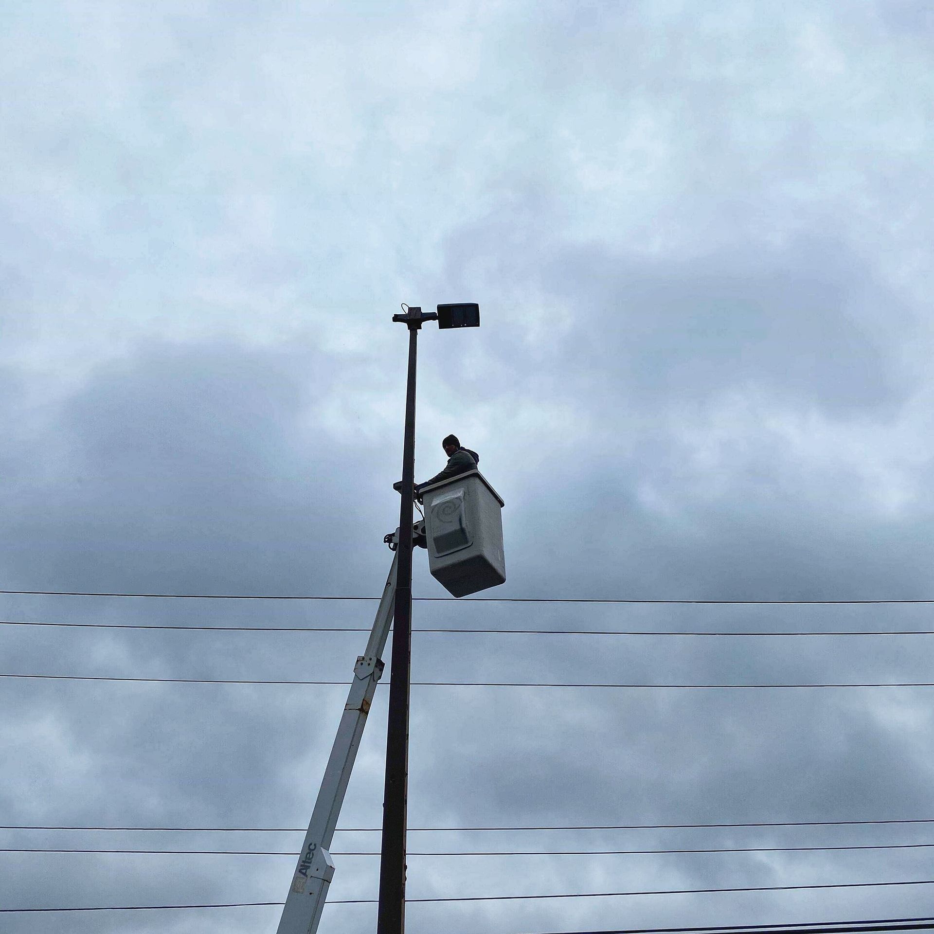 Utility worker in a lift bucket, repairing a street light on a pole, cloudy sky.