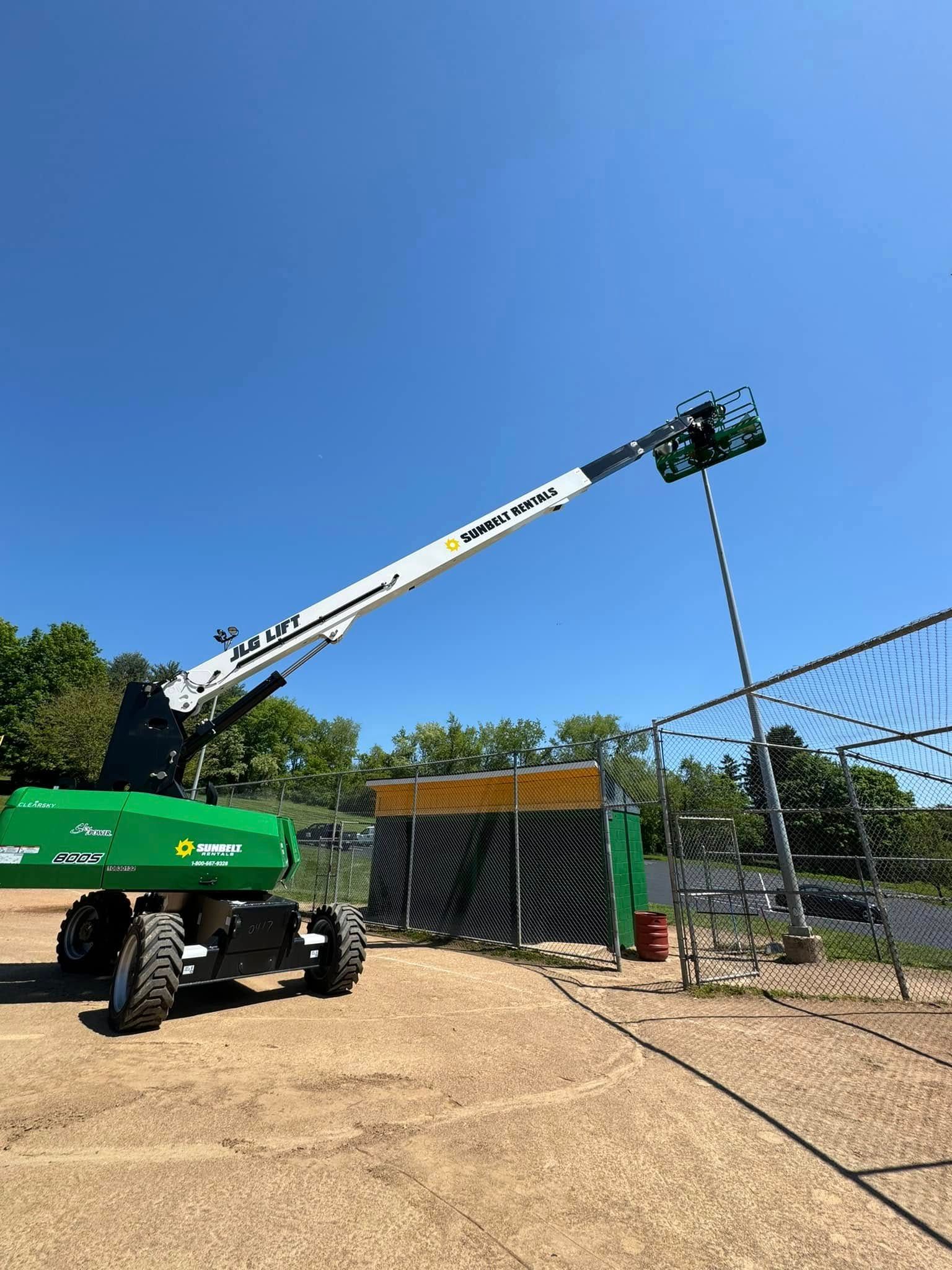 Green and white boom lift reaching towards a soccer net under a blue sky.
