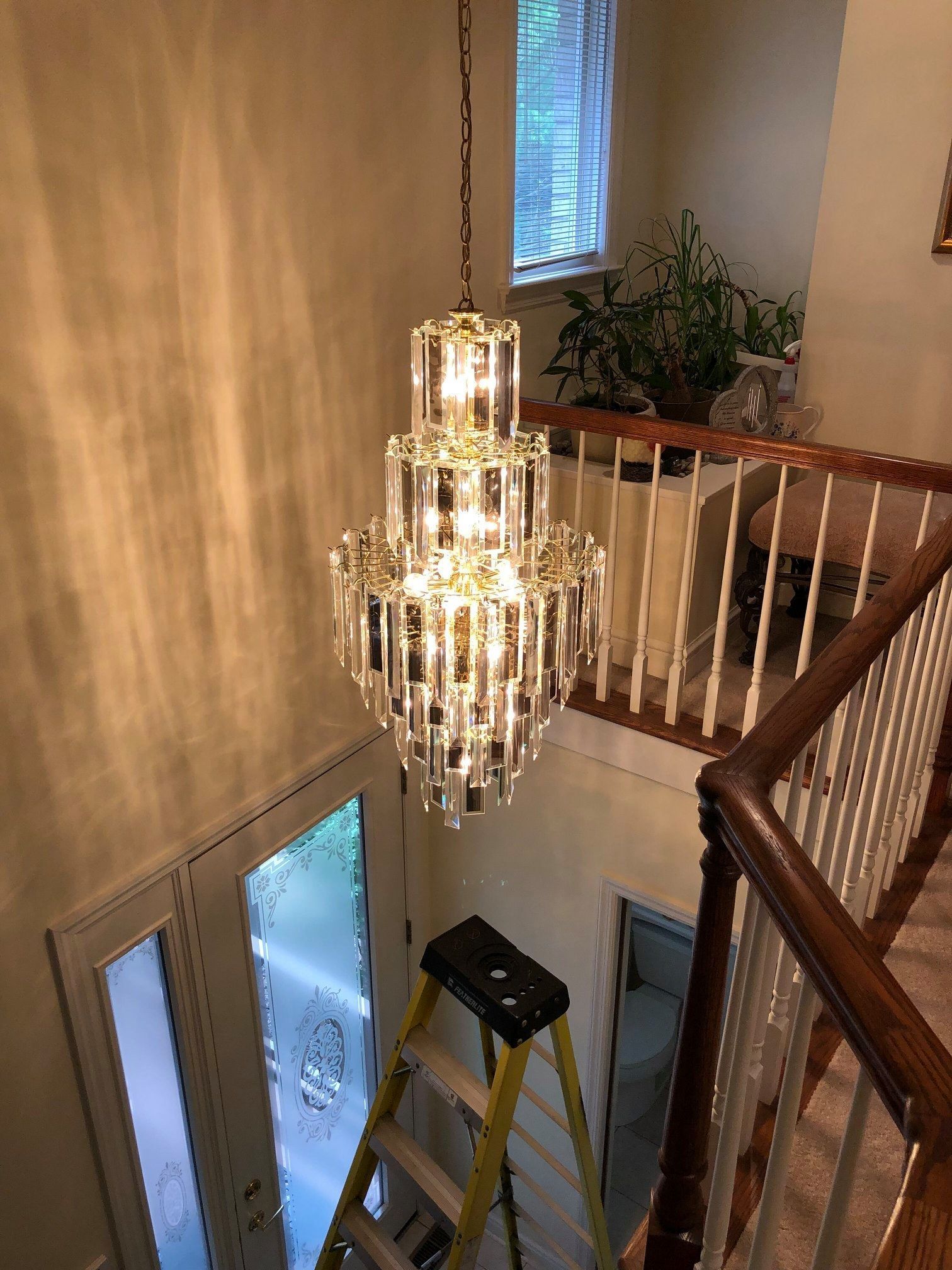 Crystal chandelier hanging above a staircase. A stepladder sits below, with a door and window visible.