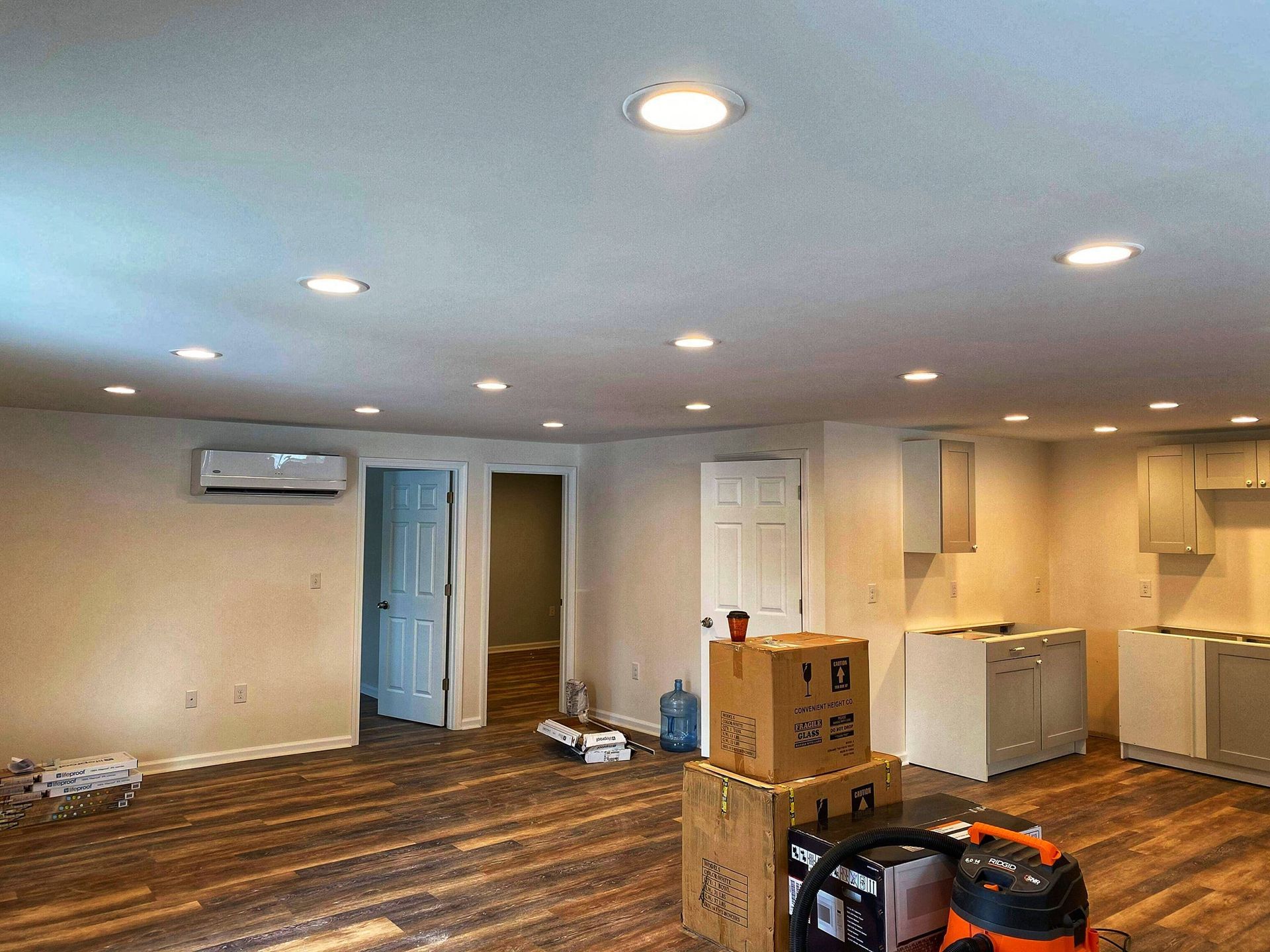 Empty room with wood flooring, recessed lighting, air conditioning unit, boxes, and unfinished cabinets.