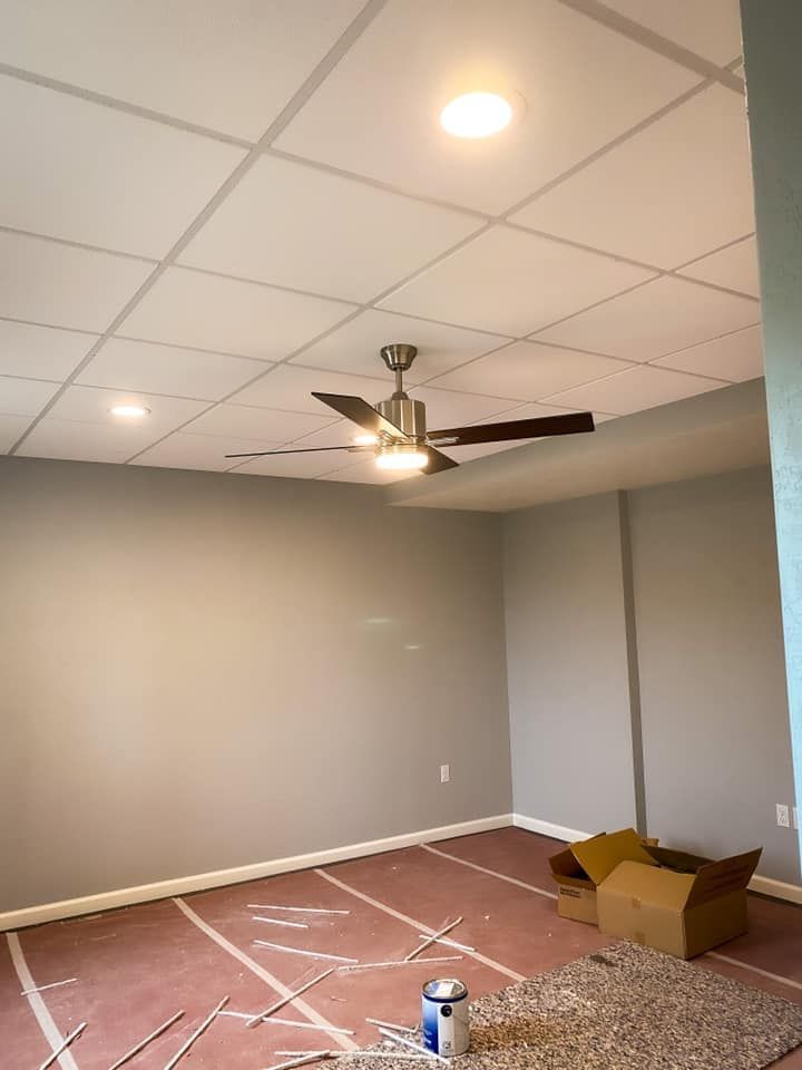 Empty room with gray walls, white ceiling tiles, ceiling fan, and exposed subfloor; construction in progress.
