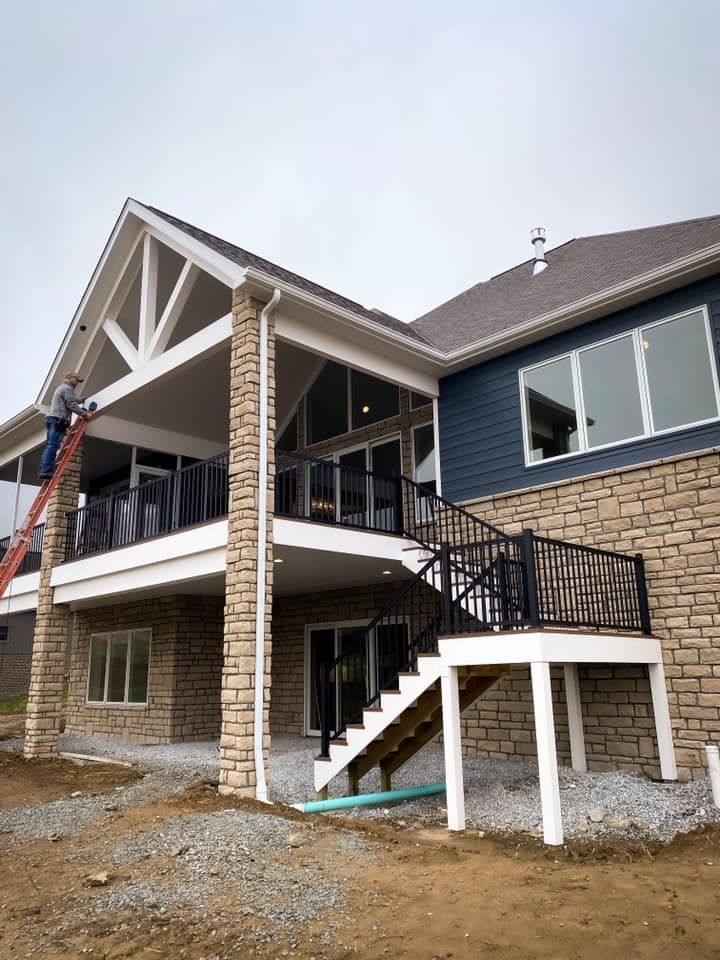 Two-story house with stone facade, covered patio, deck, and worker on a ladder.