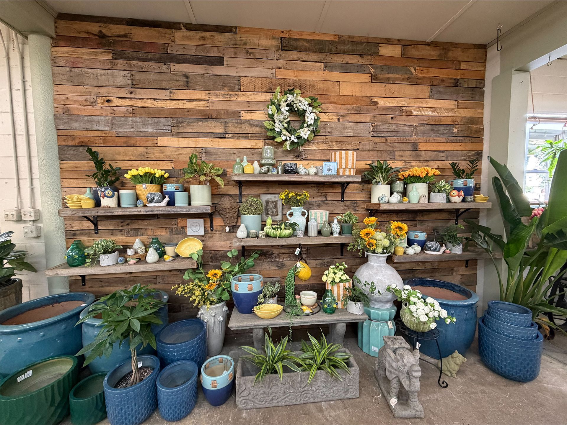 Floral shop display with potted plants, wreaths, and decorative items on shelves against a wood wall
