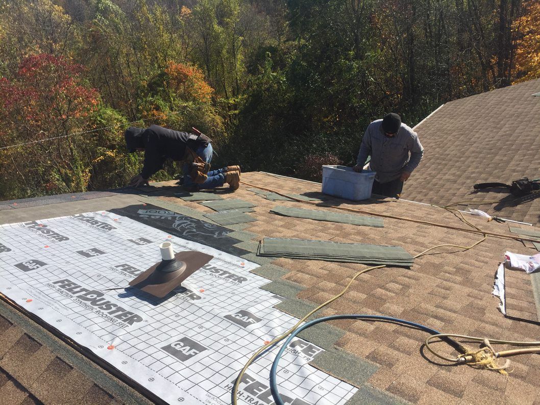 Roof workers installing shingles with tools and materials on a sunny rooftop, surrounded by trees.