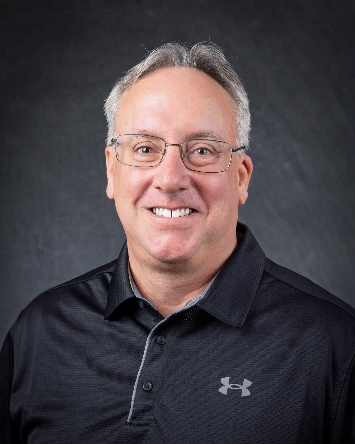 Man with glasses, smiling, wearing black shirt with gray logo, in front of a gray backdrop.