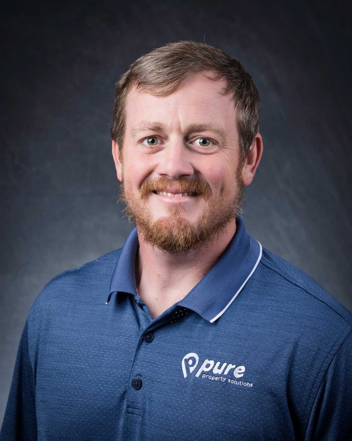 Man with auburn beard in a blue polo shirt, smiling, against a dark background, logo on shirt.