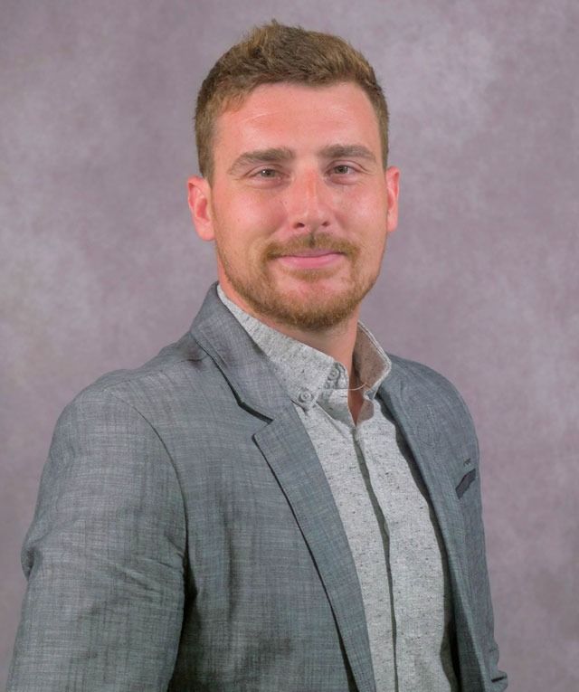 Man in gray blazer and button-down shirt smiles, looking at the camera. Neutral background.
