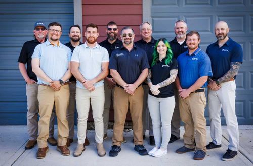 Group of 12 people smiling in front of a building. Several are wearing company shirts,.