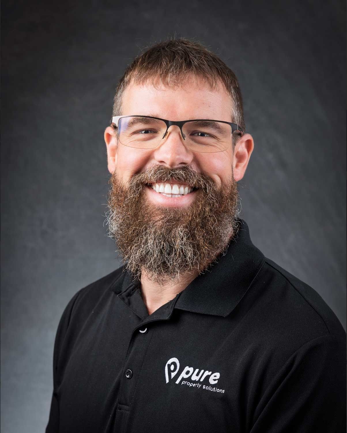 Man with a beard, glasses, and black polo shirt smiles, posing for a portrait. Background is gray.