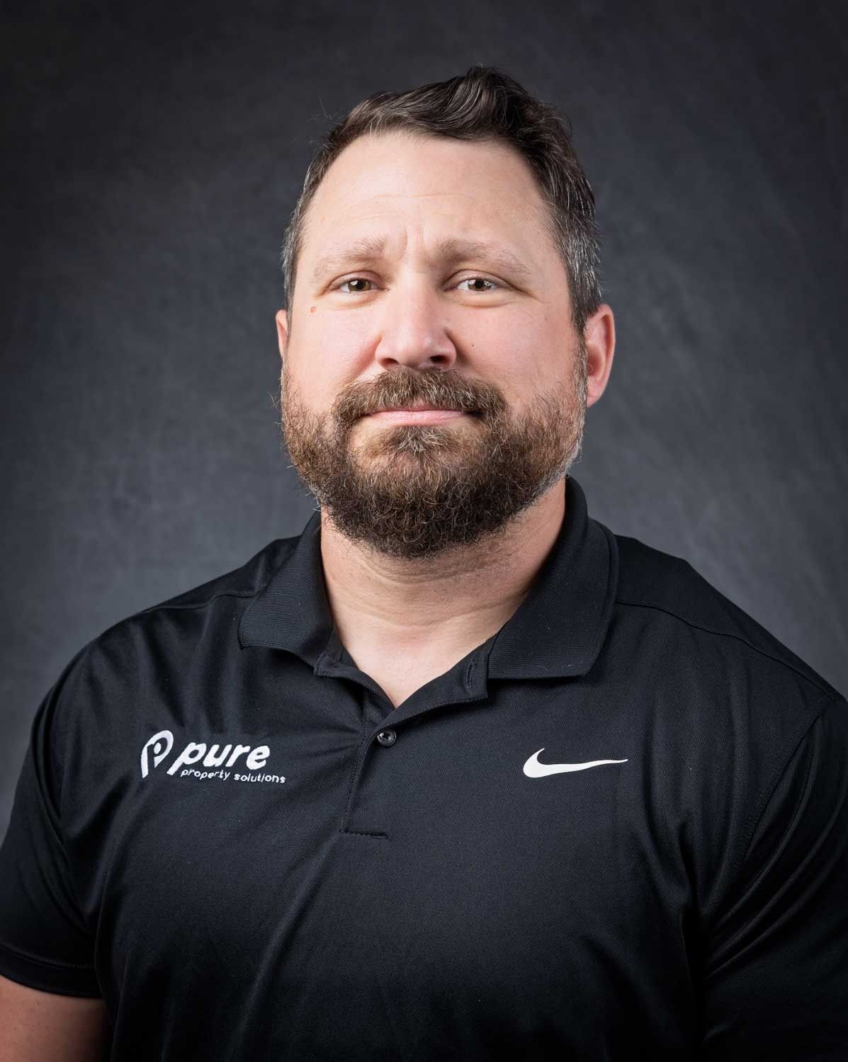 Man with a beard in a black polo shirt, logo on chest, Nike swoosh. Dark background.