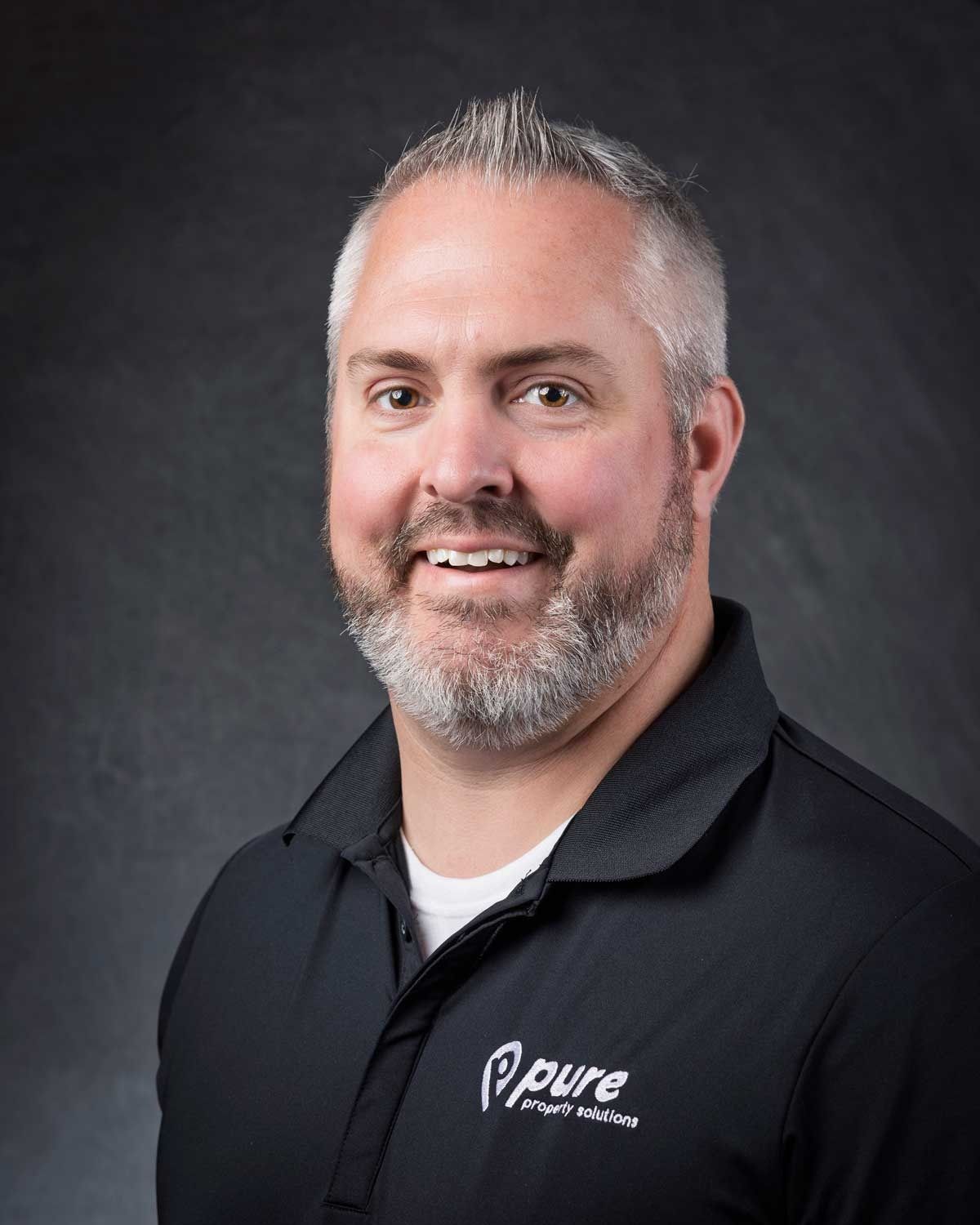 Man with graying hair and beard smiling, wearing a black polo shirt with 