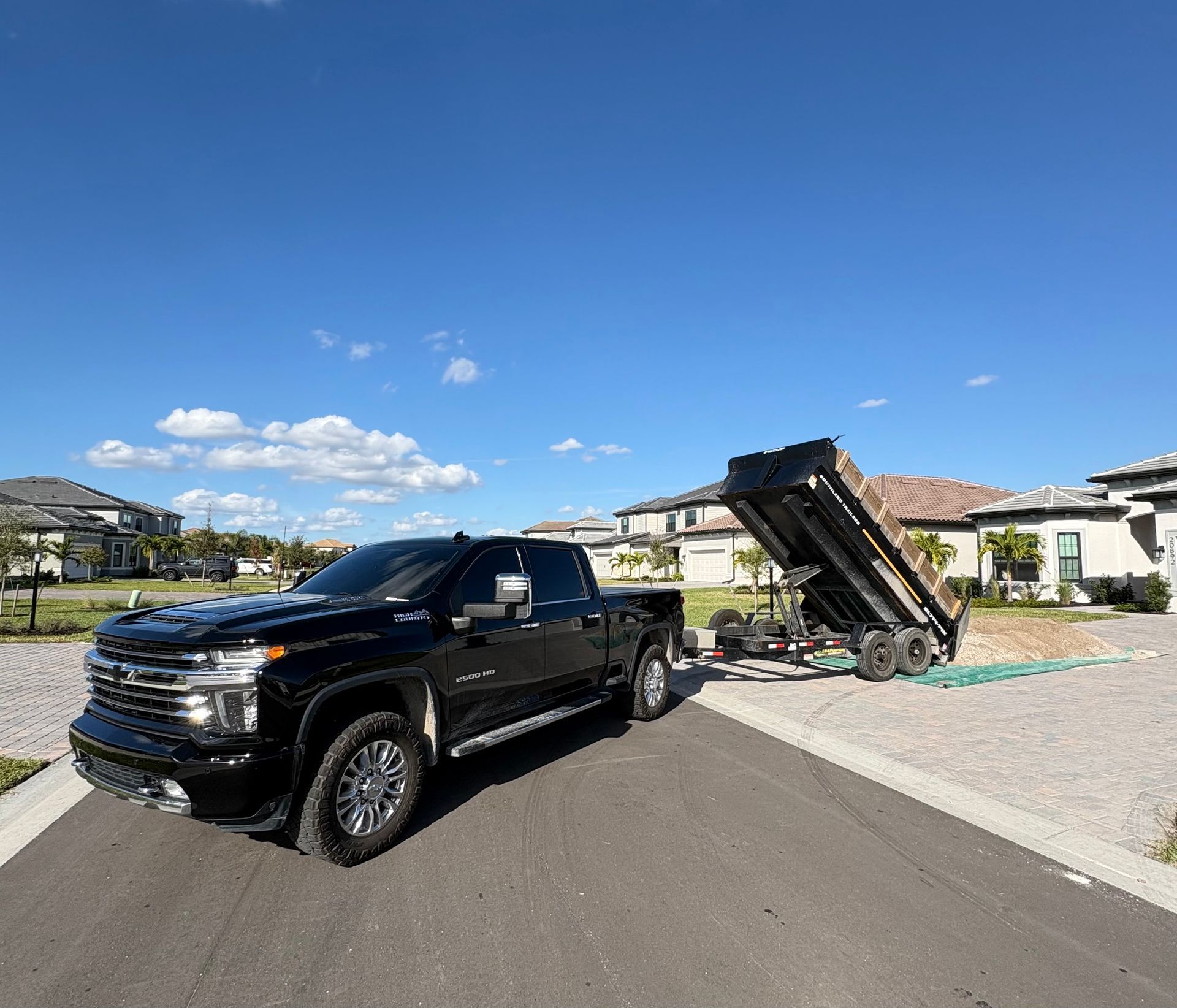 A black pickup truck towing a dump trailer on a driveway. Clear blue sky, suburban setting.