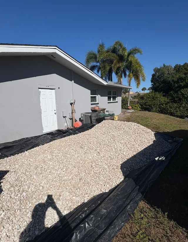 A gravel path lay against a gray building with a white door. Black landscape fabric visible, blue sky.