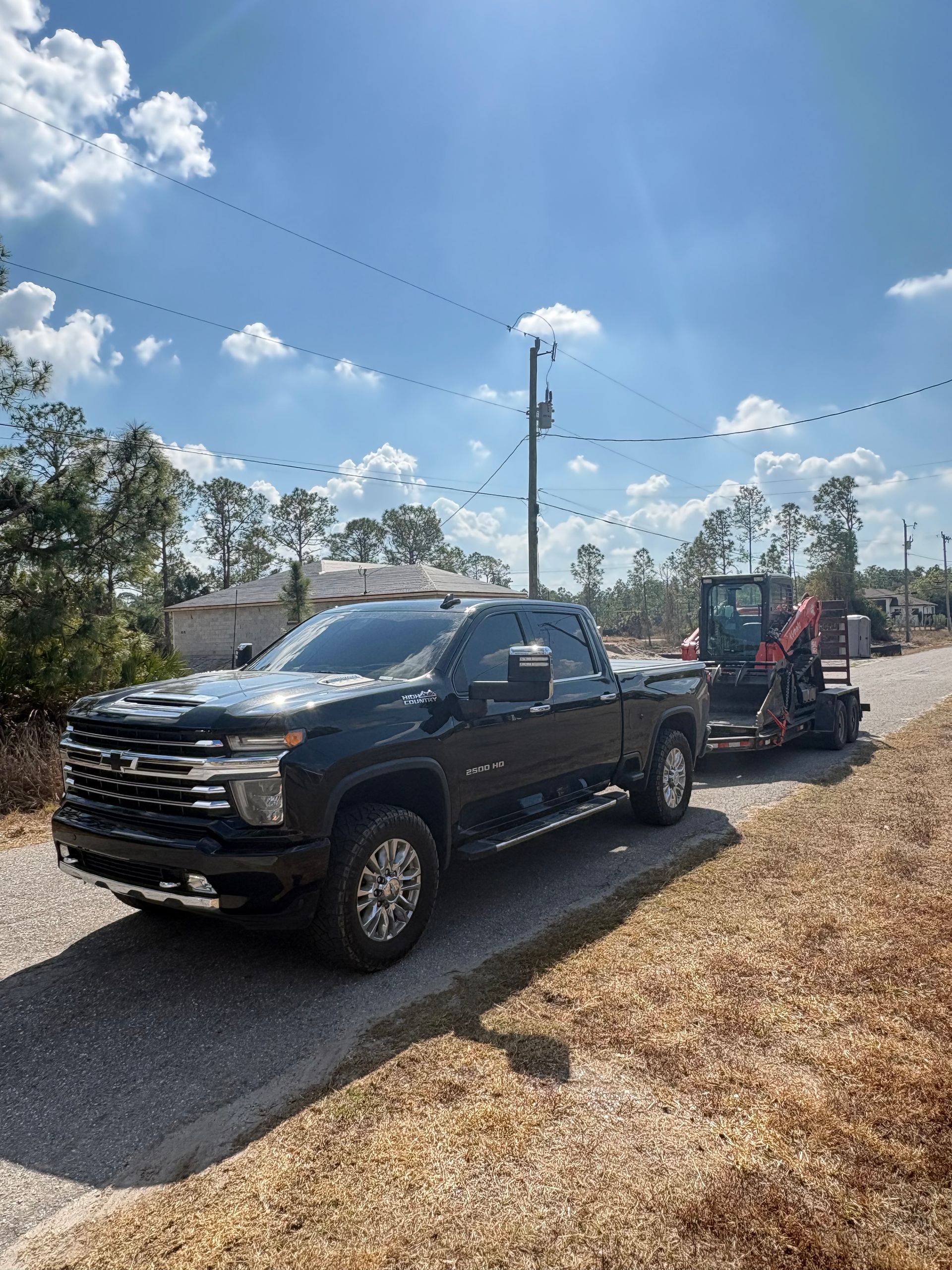 A black pickup truck towing a mini excavator on a trailer parked on a gravel road under a sunny sky.