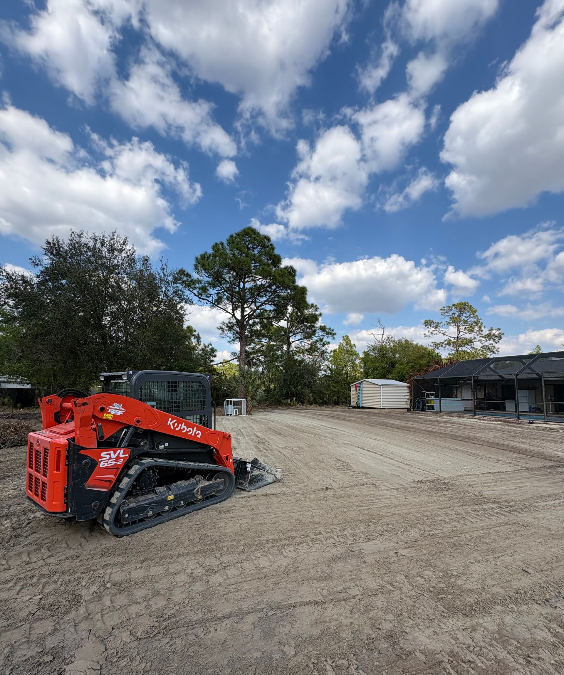 Red track loader on a dirt lot under a cloudy blue sky, near trees and a building.