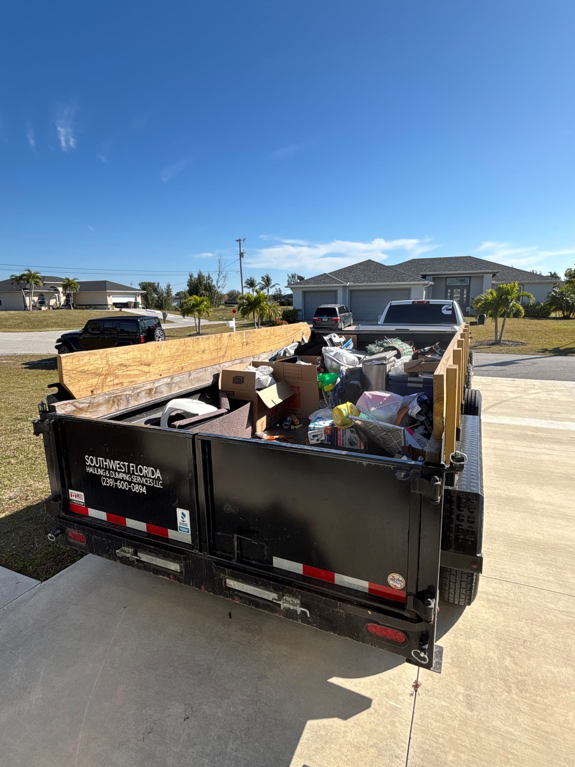 A black trailer filled with trash, parked in a driveway on a sunny day. Houses and a truck are in the background.