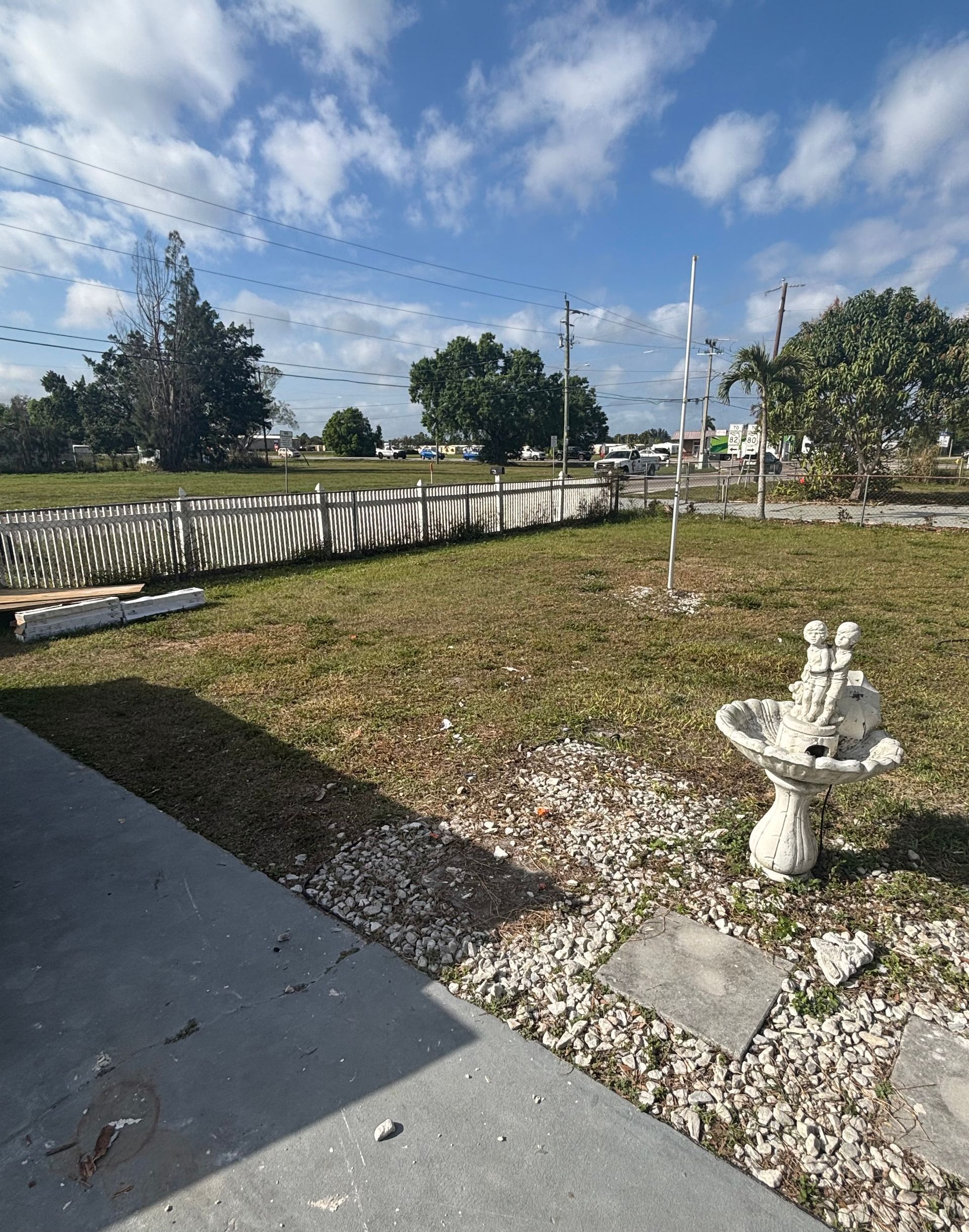 A sunny lawn with a white fence and a small birdbath, blue sky with clouds.