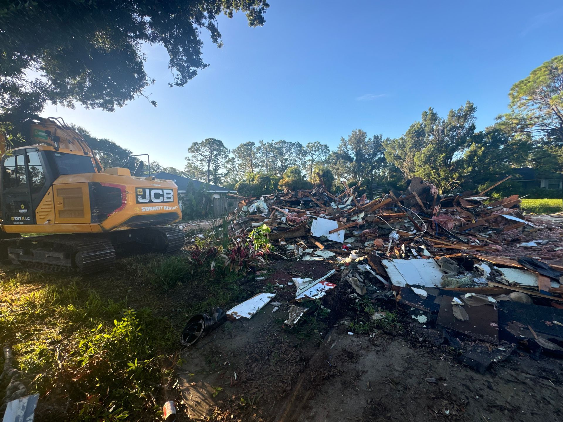 Yellow excavator demolishing a pile of debris, likely a building. Green grass and trees under a blue sky.