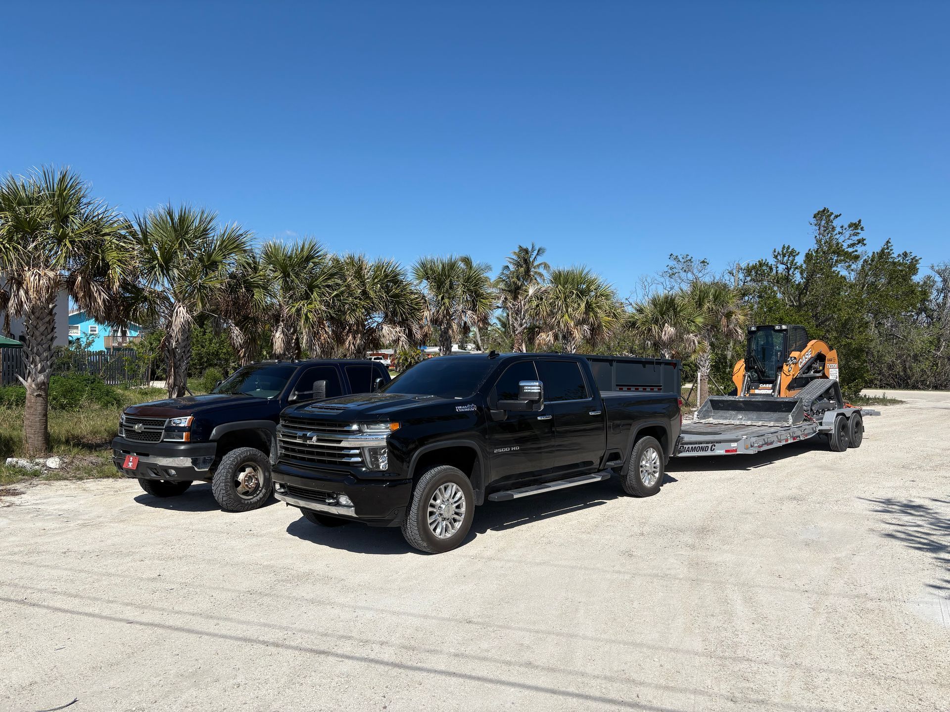 Two black pickup trucks parked on gravel; one towing a trailer with a small excavator.