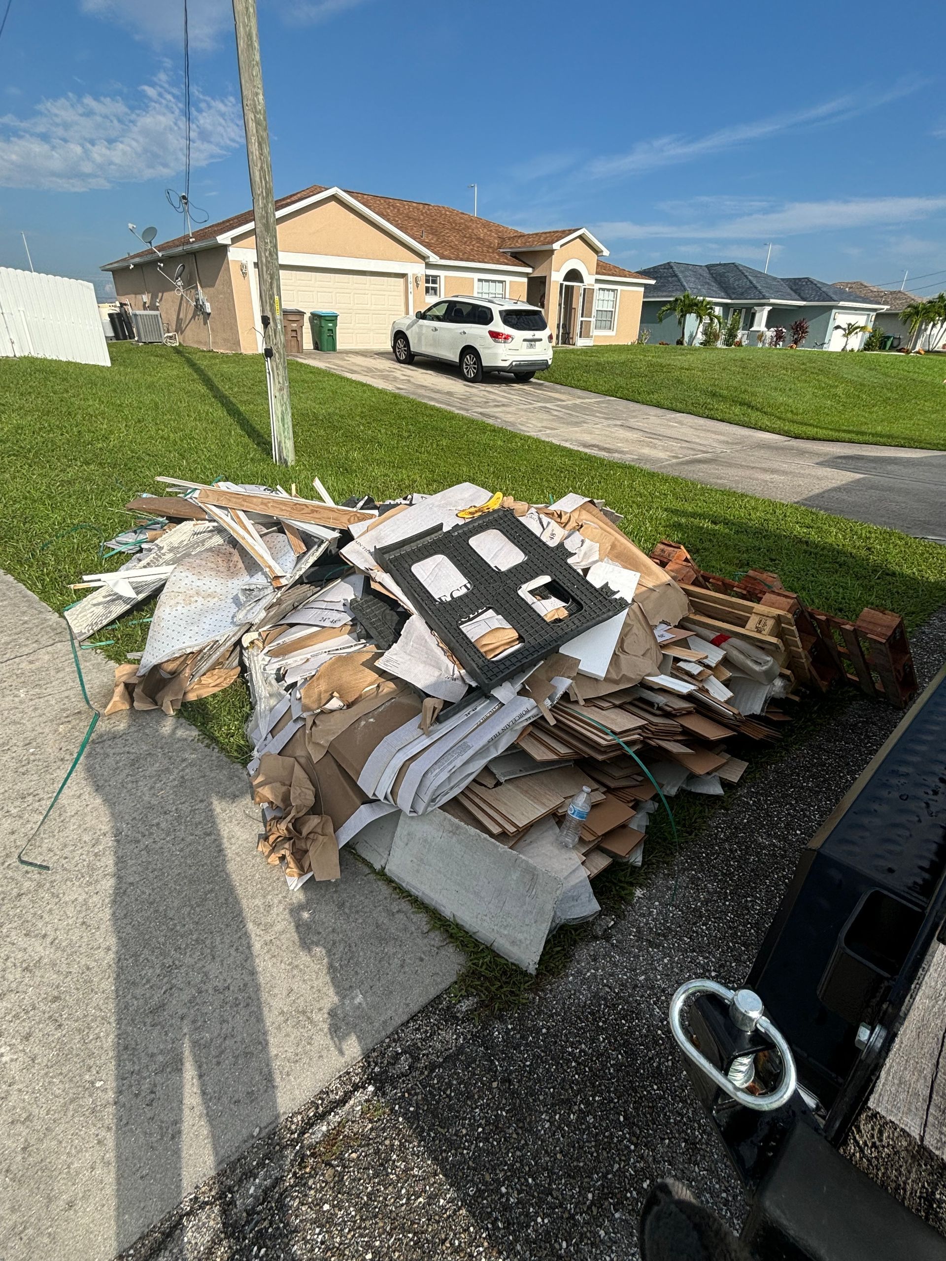 Pile of discarded cardboard, wood, and debris on a sidewalk near a residential house and parked car.