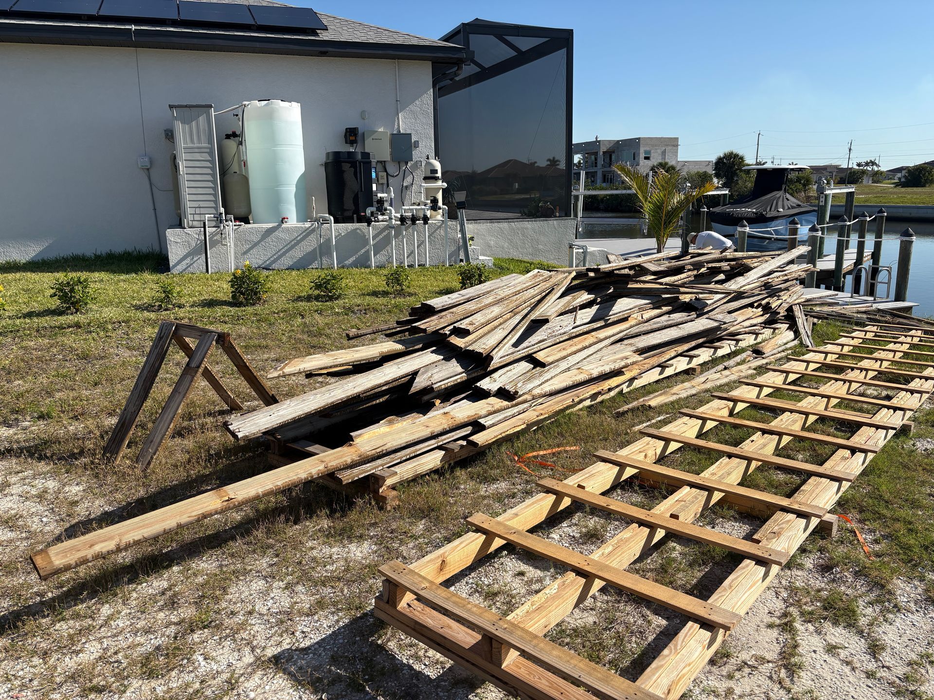 Pile of weathered wooden planks and a wooden ladder on a grassy yard near a canal and a house.