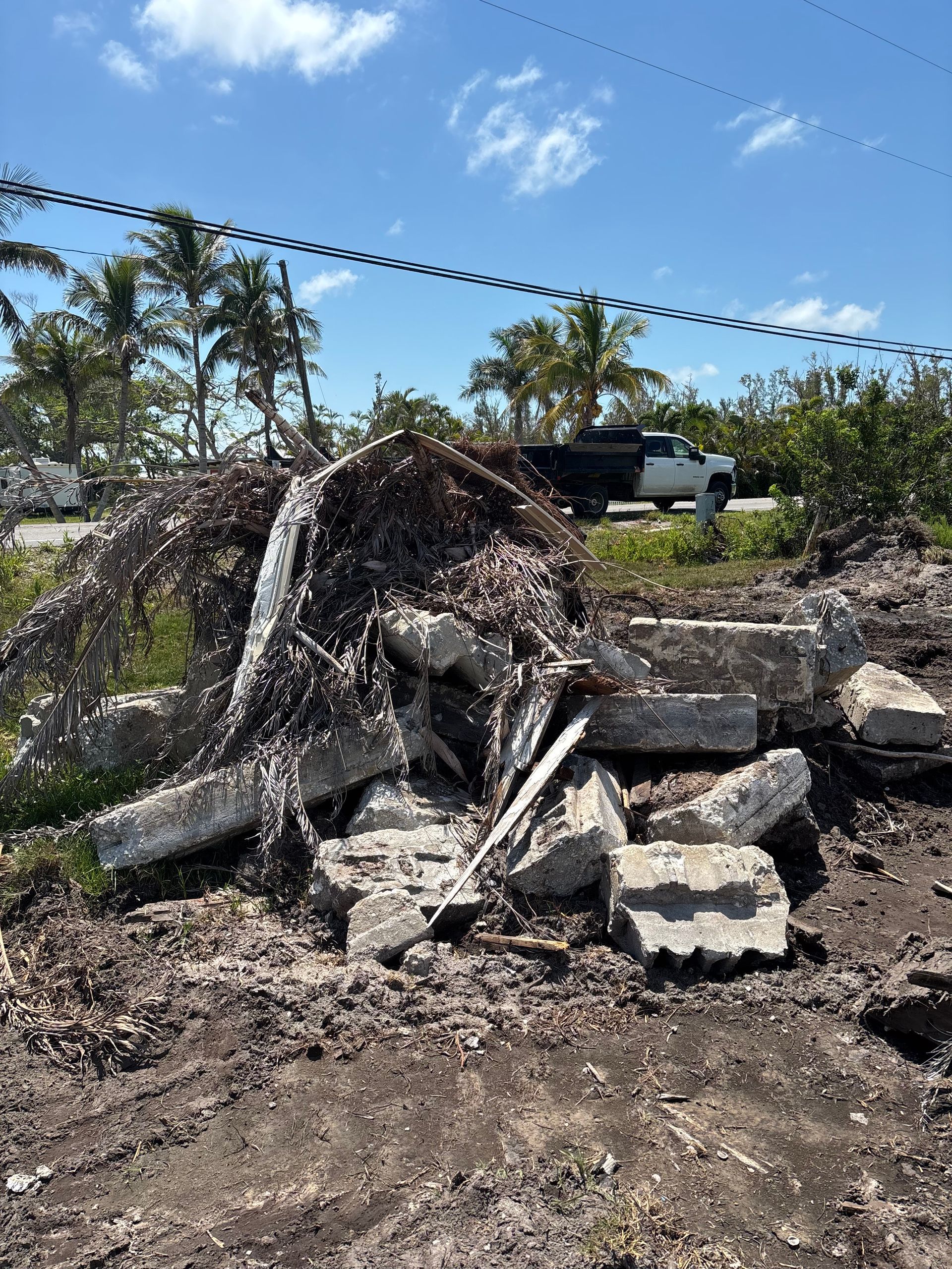 Debris pile of broken concrete and vegetation on dirt ground; truck in background under blue sky.