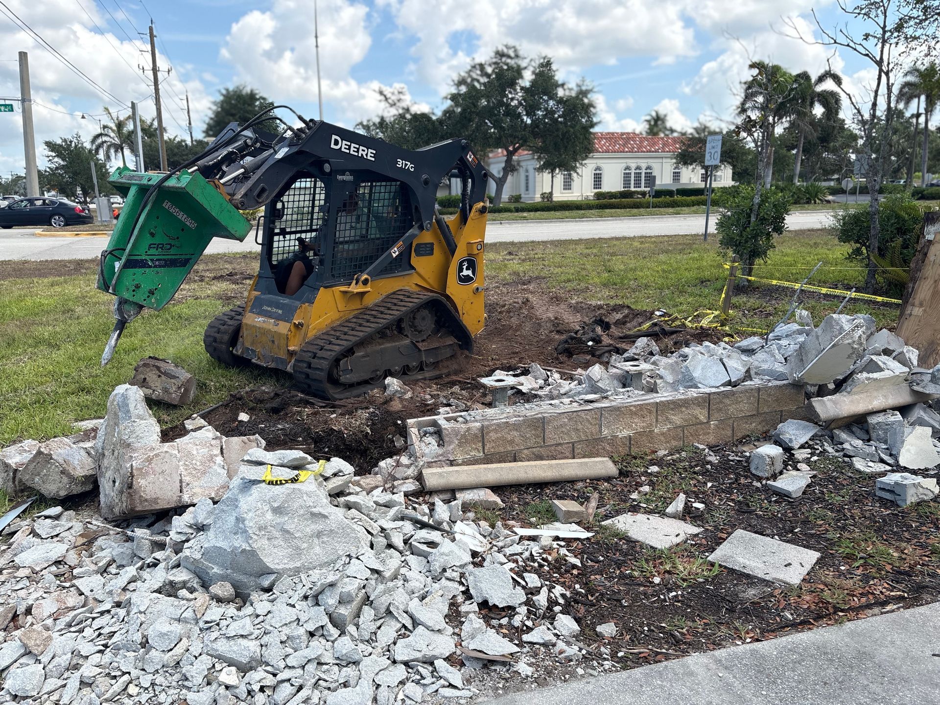 A yellow skid steer with a jackhammer attachment demolishes a concrete wall in a grassy area.