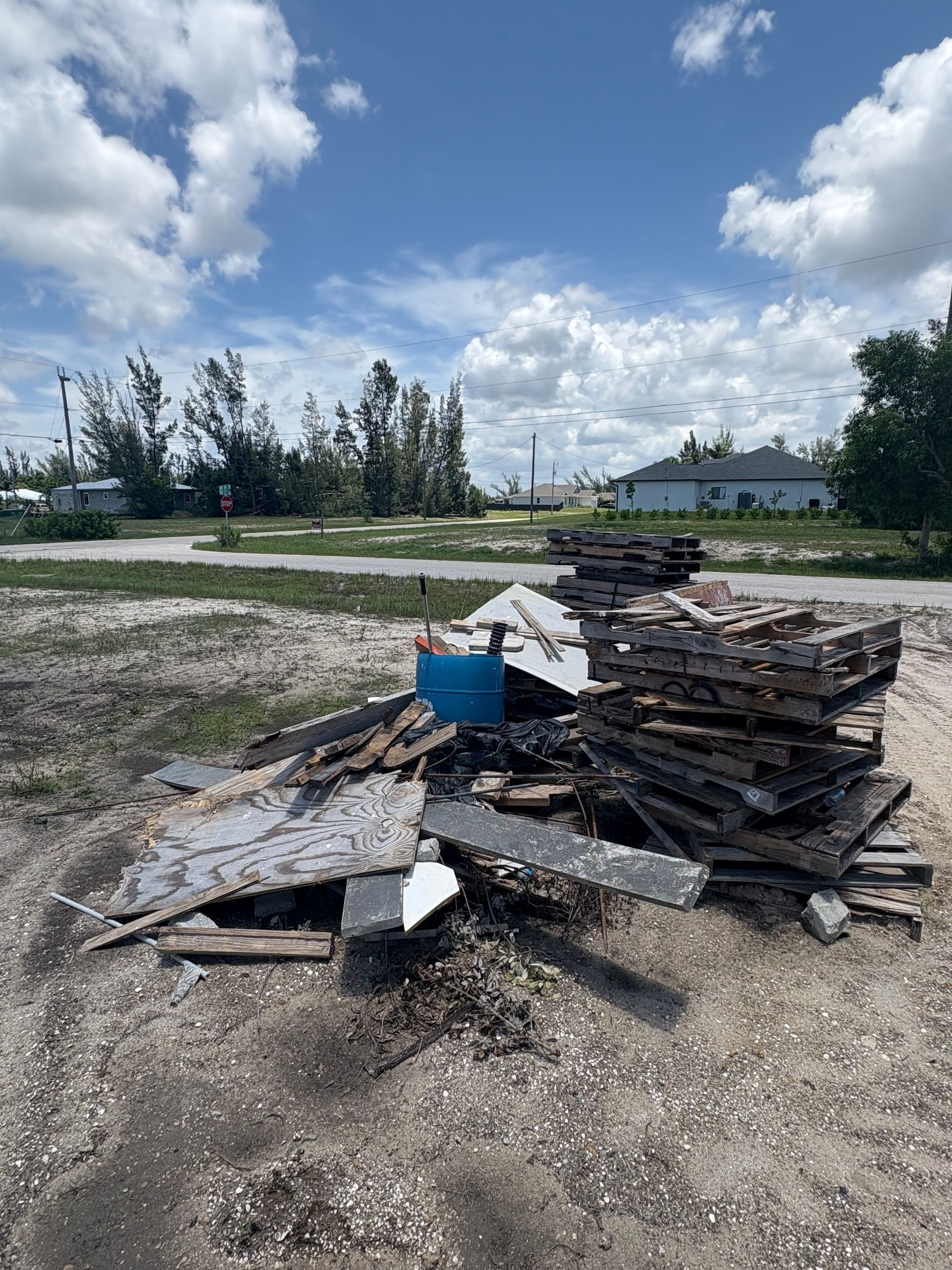 Pile of debris, possibly from a demolished building, on a dirt patch with a blue bin. Blue sky, clouds, and trees in background.