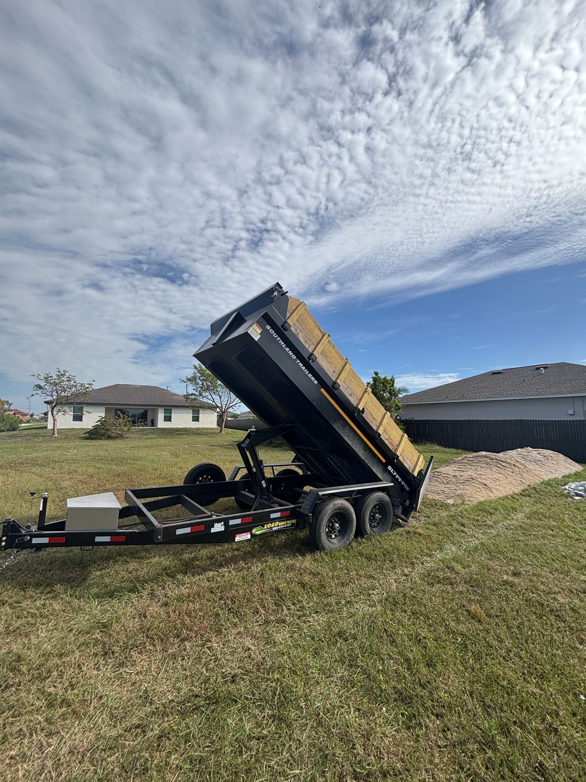 A black dump trailer tilted in a grassy yard under a partly cloudy blue sky.