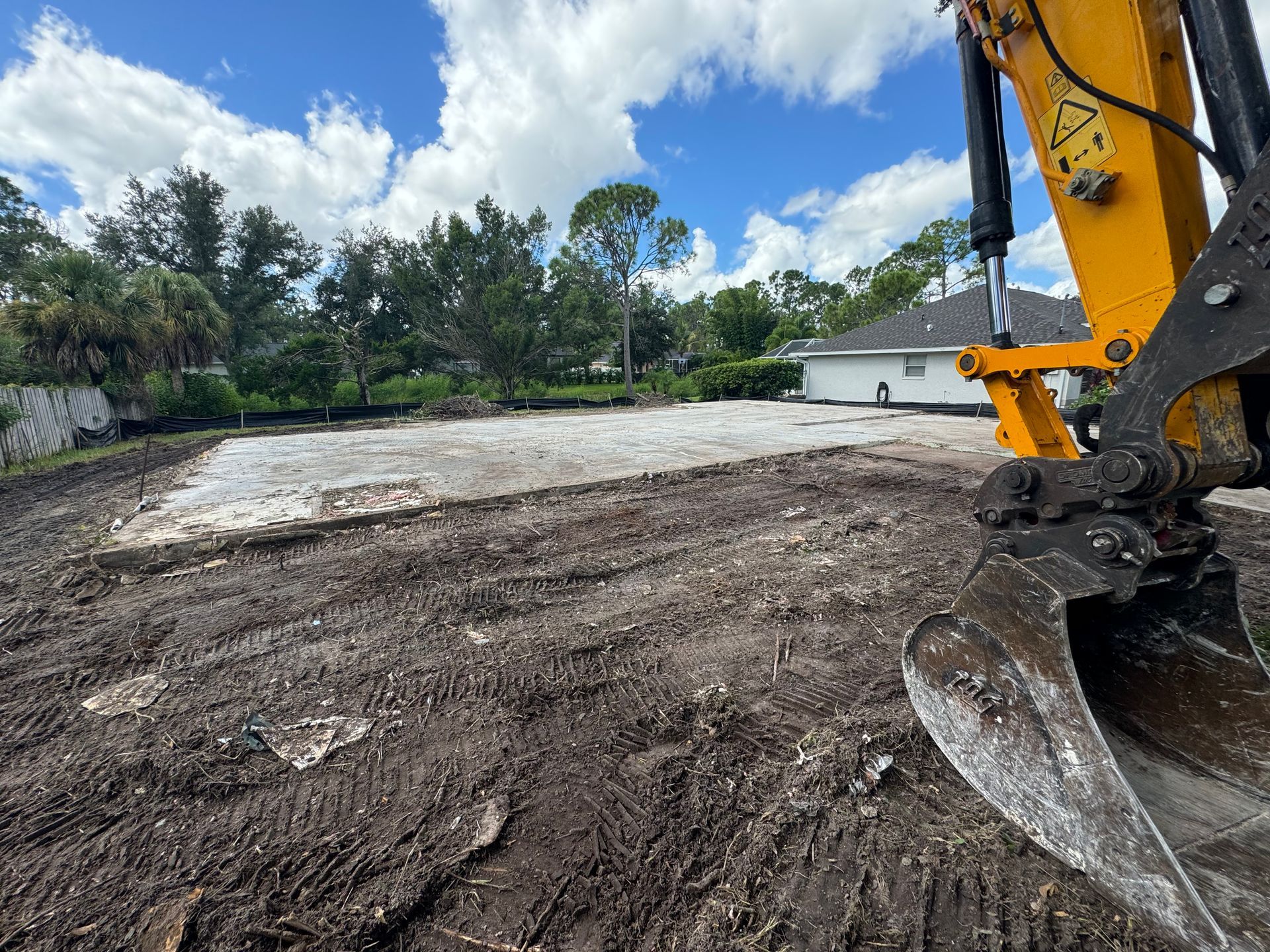 Excavator and freshly cleared land with a white house in the background on a sunny day.