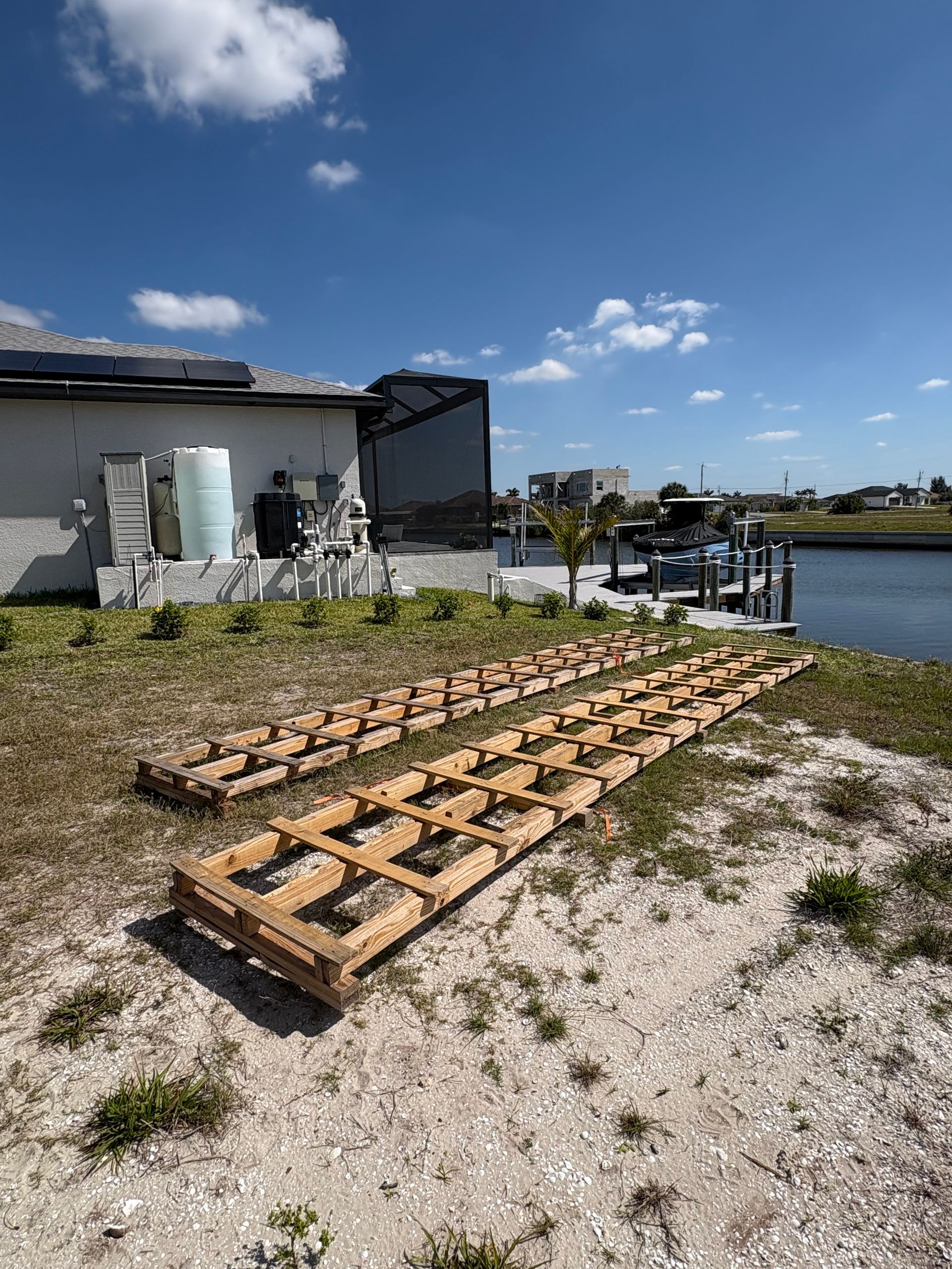 Two wooden frames on a sandy yard near a waterfront home on a sunny day.