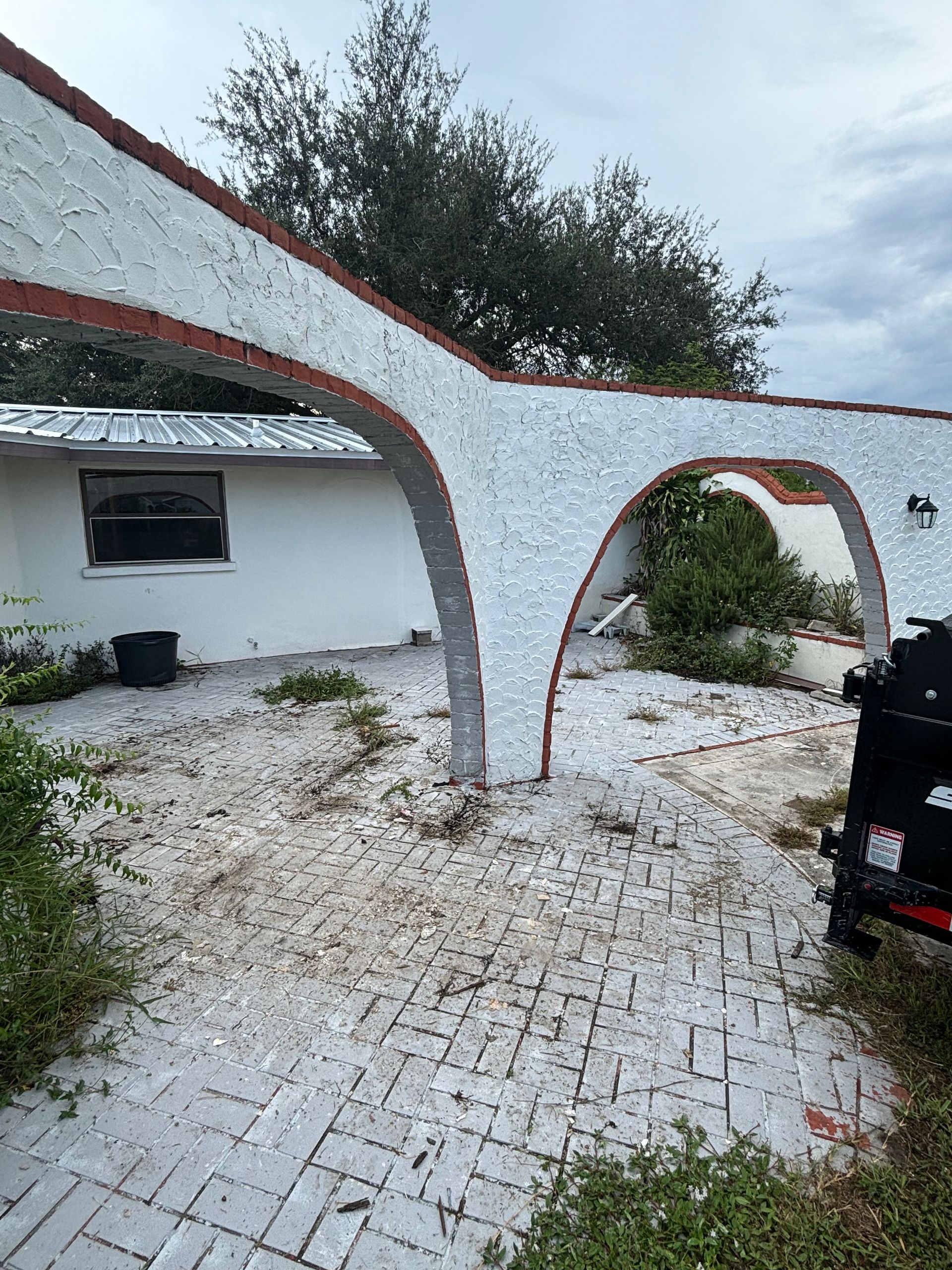 Exterior of a white stucco building with arched structures, brick pavers, and some weeds; overcast sky.