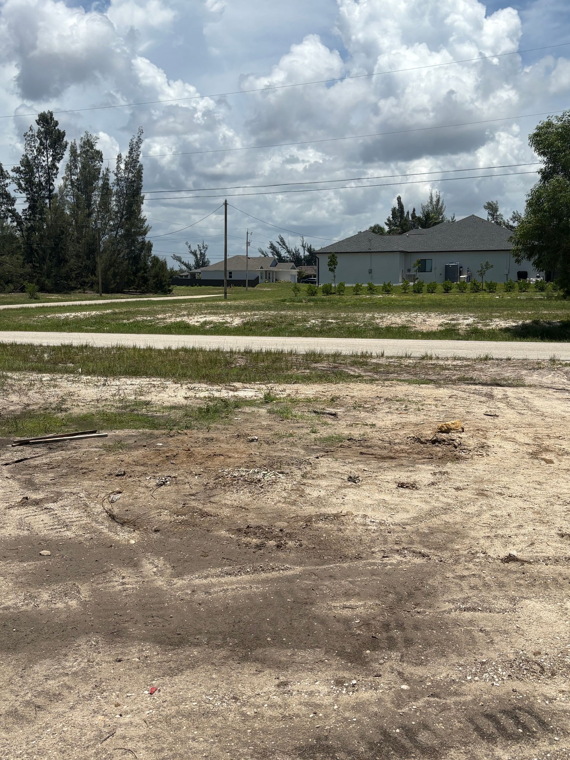 Dirt road leading to a grassy lot with houses and trees under a cloudy sky.
