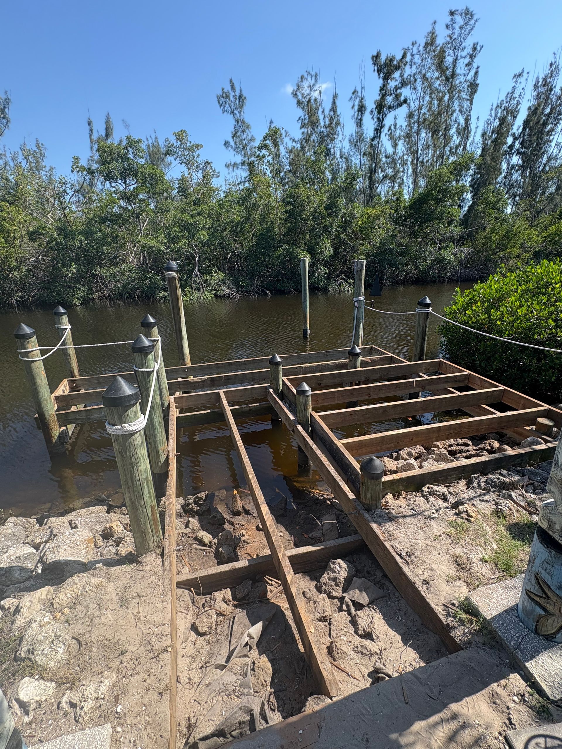 Dock partially dismantled over water, exposed beams, pilings, and dirt, with vegetation in background.