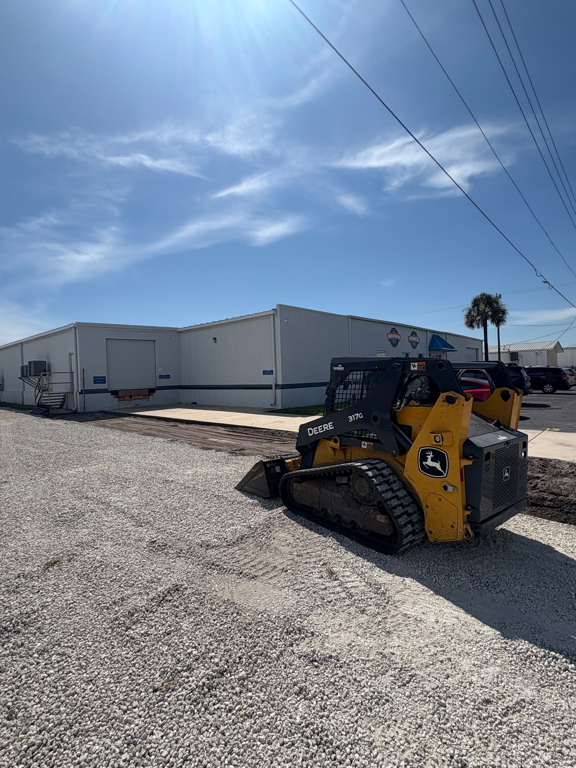 Yellow and black John Deere skid steer moving gravel in front of a white industrial building under a blue sky.