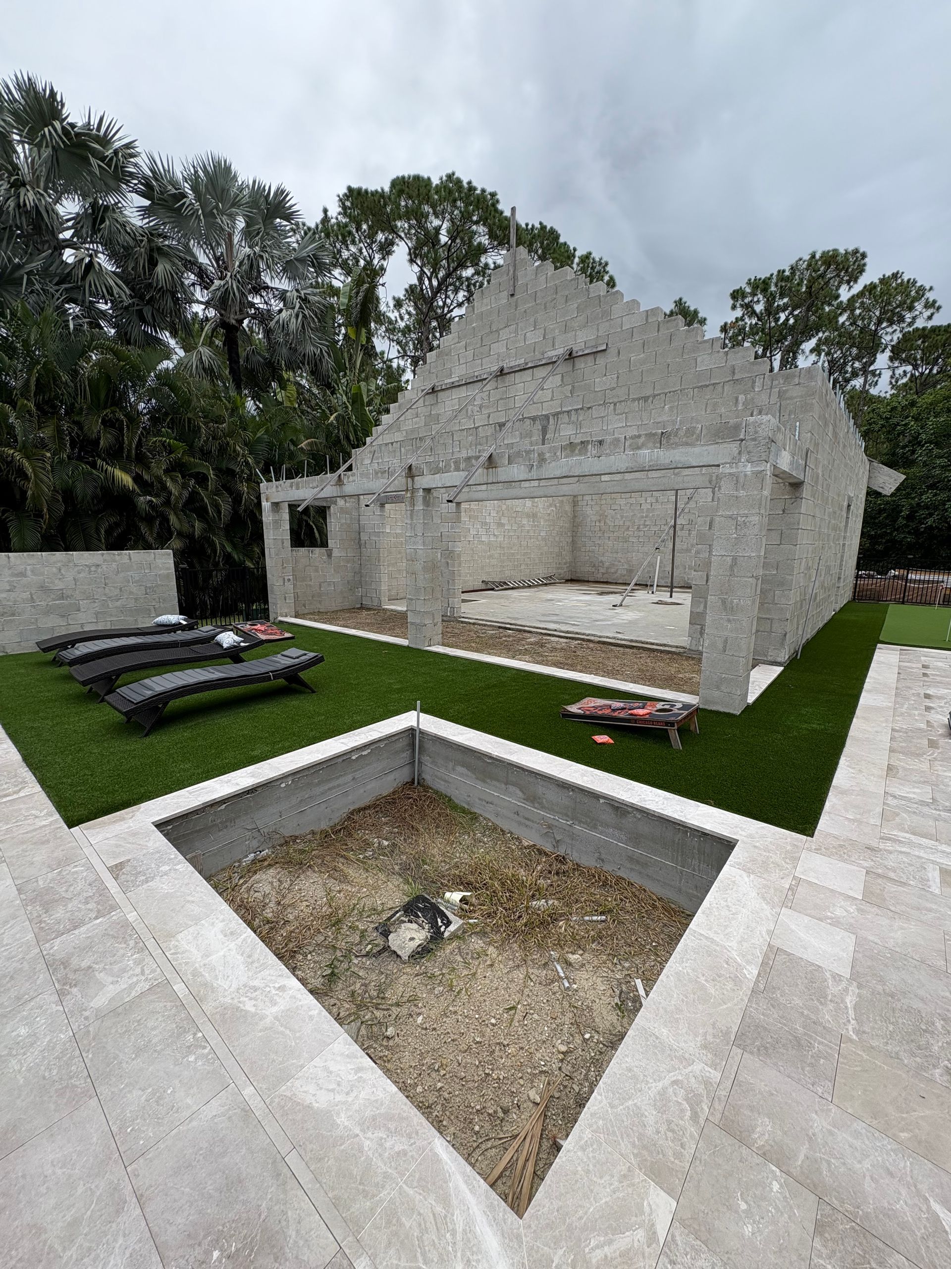 Stone building under construction with a grassy yard and square stone-lined pit.