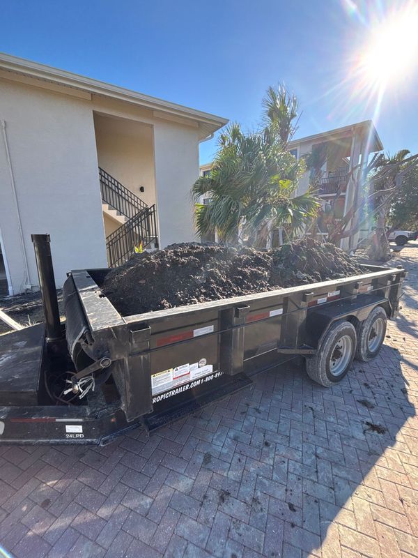 Black dump trailer filled with dark soil parked on a brick-paved surface near a light-colored building.