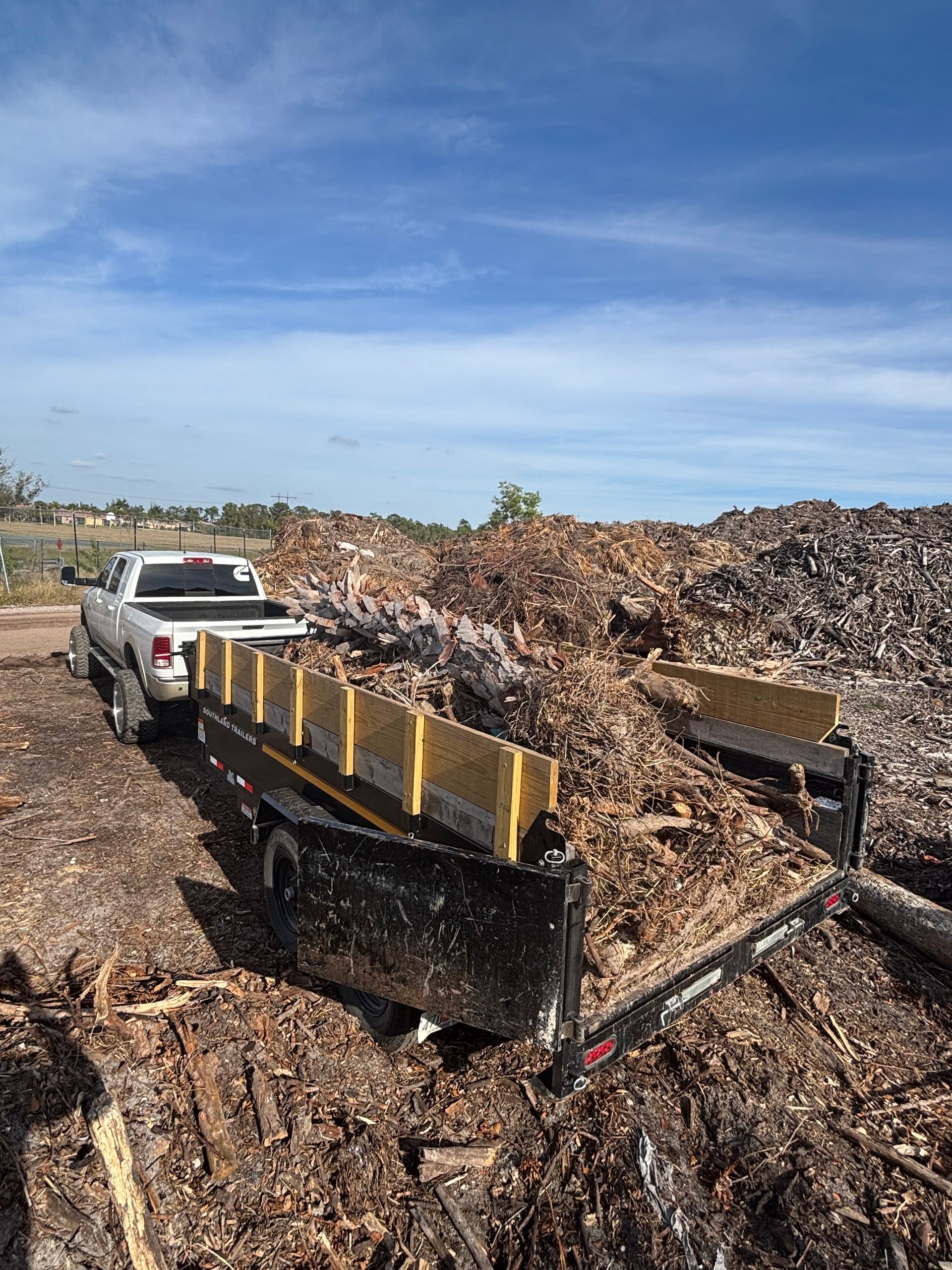 Truck with trailer being loaded with wood chips at a large pile outdoors under a blue sky.