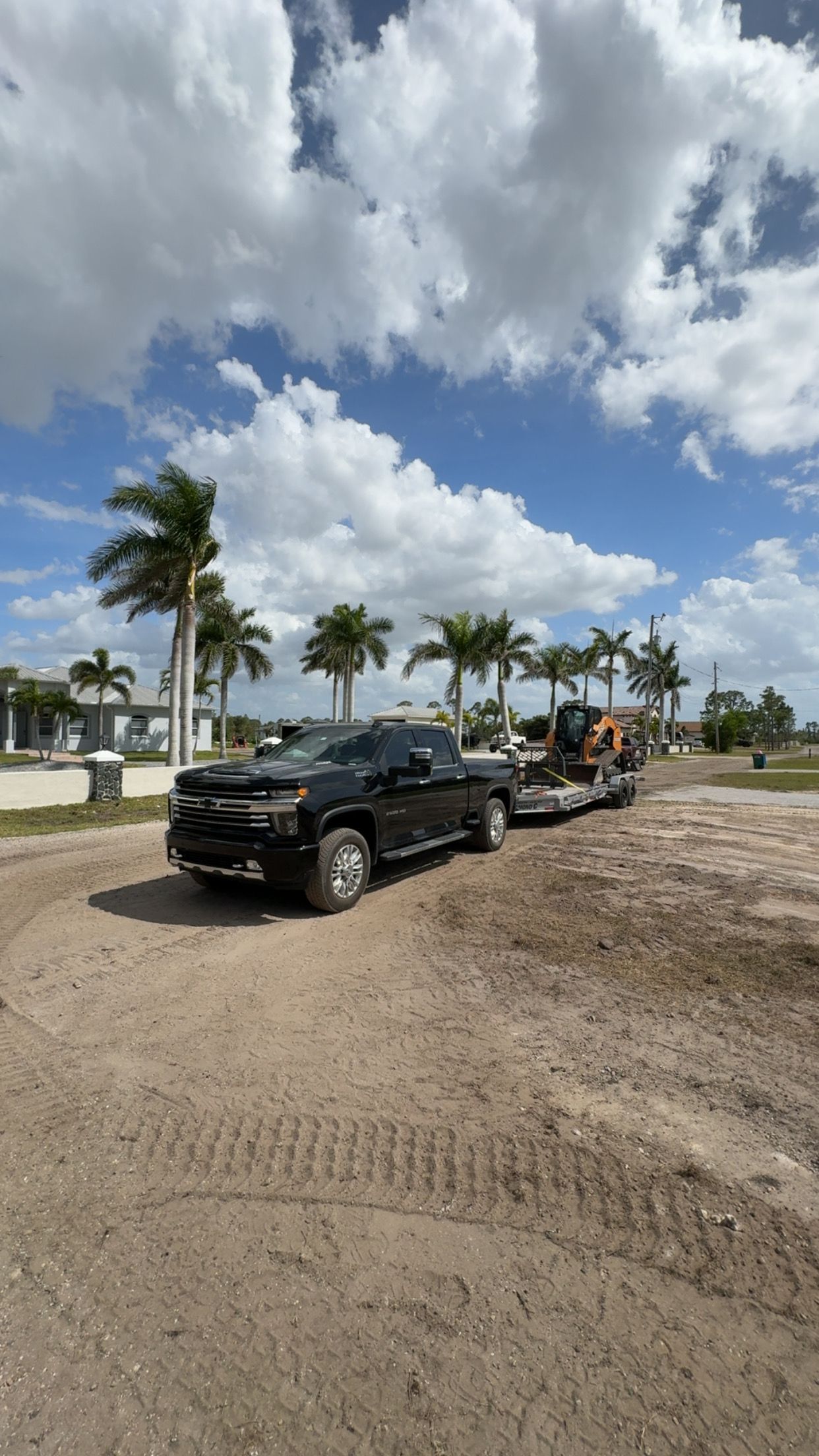 Black truck towing orange construction equipment on a dirt road, palm trees, sunny sky.