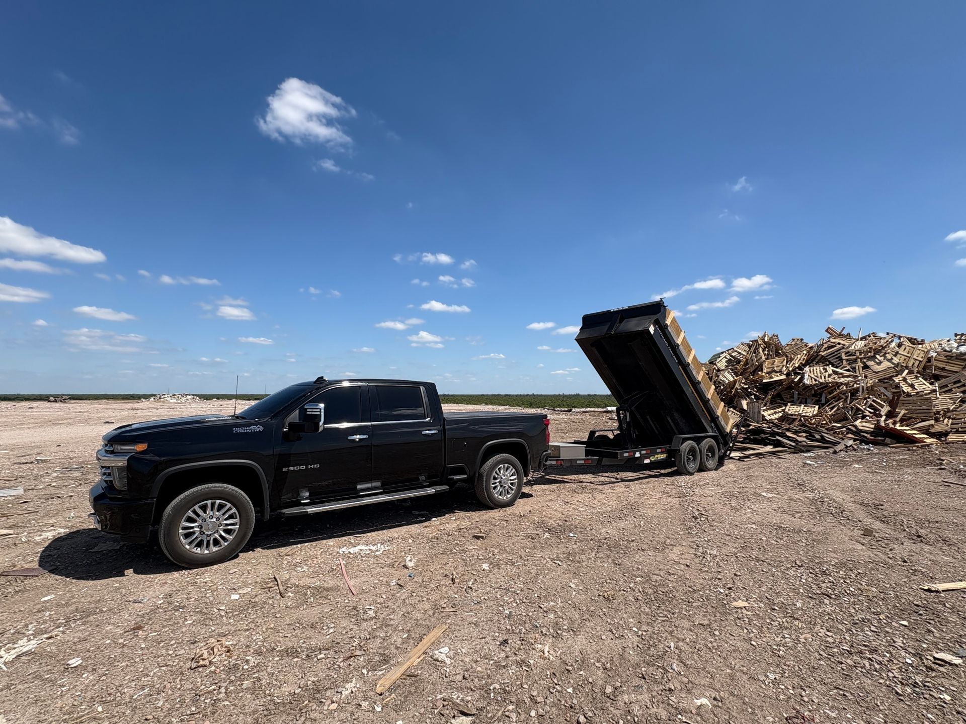 Black truck towing a trailer filled with wood debris on a barren, sunny field.