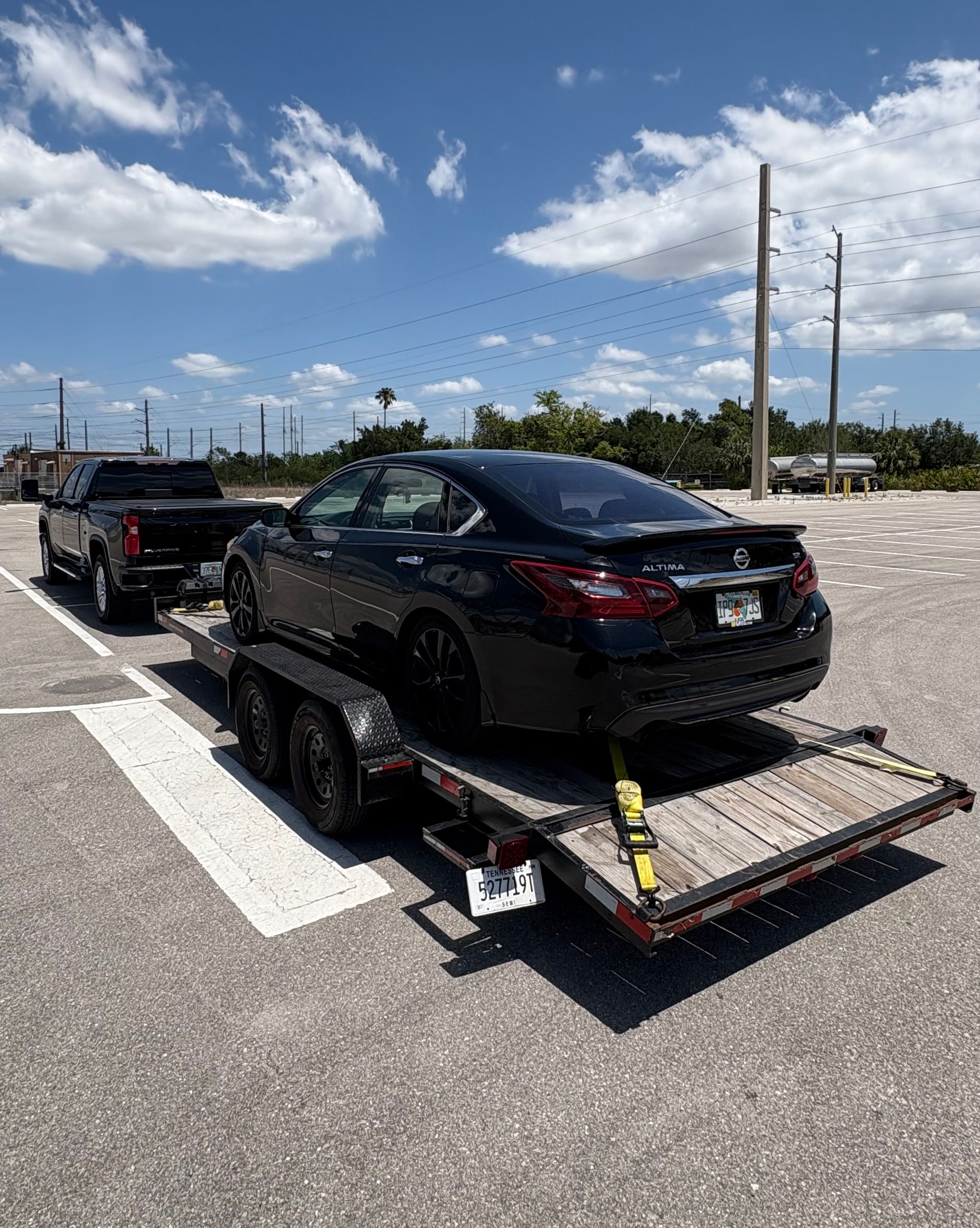 Black car on a trailer being towed by a black truck in a sunny parking lot.